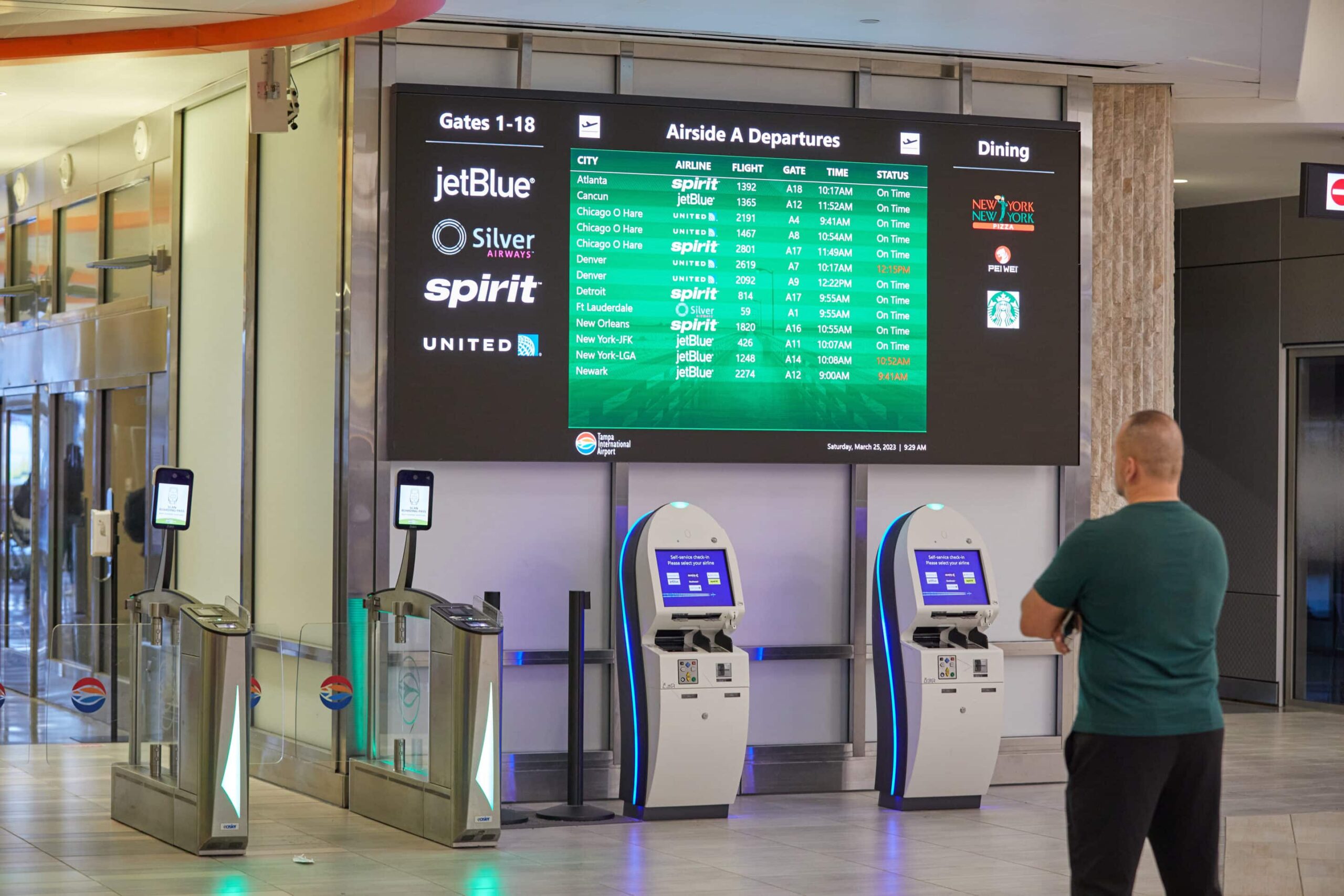 A man at an airport terminal views JetBlue, Silver, Spirit, and United flights on a digital board near self-check-in kiosks.