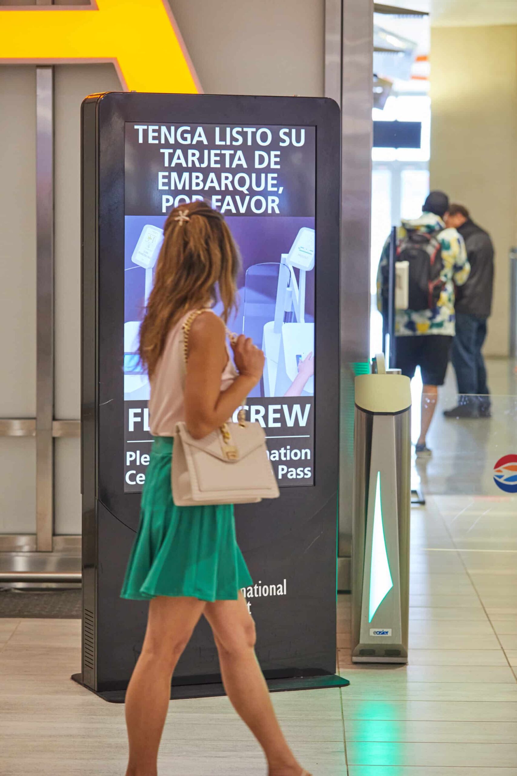 A woman in a green skirt and white top passes an airport sign about boarding passes. Other travelers are visible.