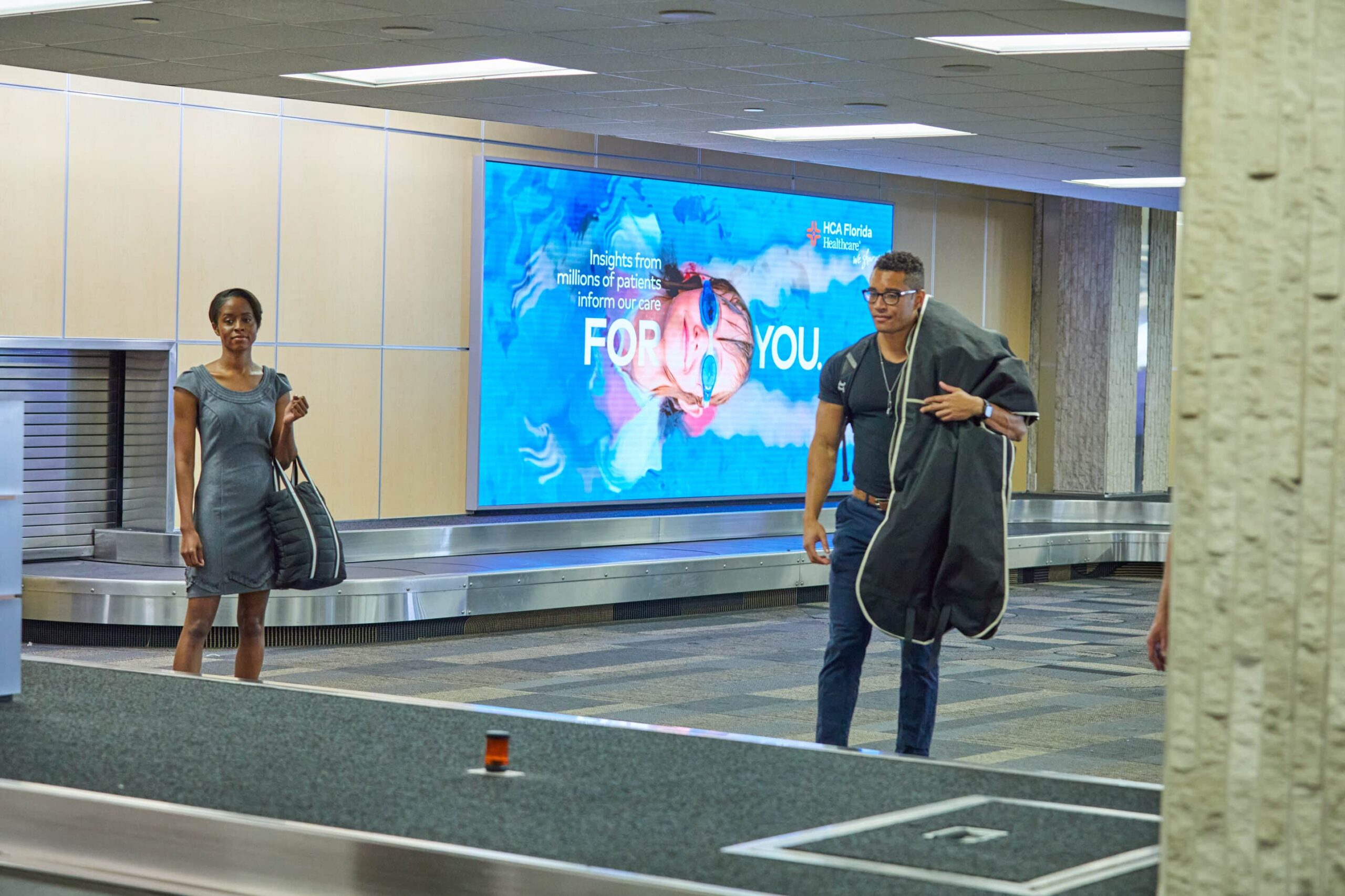 A woman with a black bag and a man with a garment bag stand by airport baggage claim, near an ad promoting healthcare services.