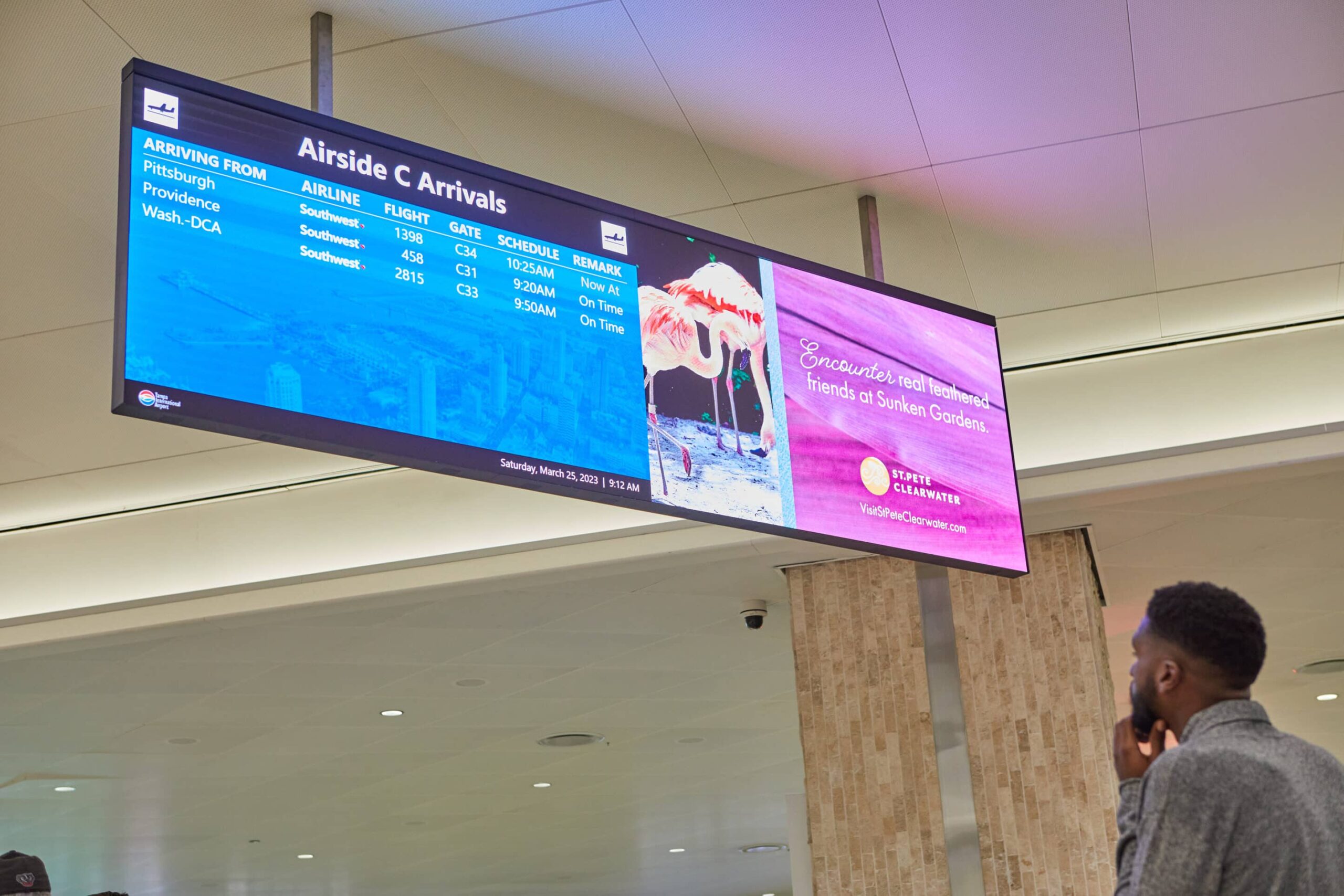 A person gazes at an airport departures board showing flights to Pittsburgh and Providence, plus an ad with pink flamingos.