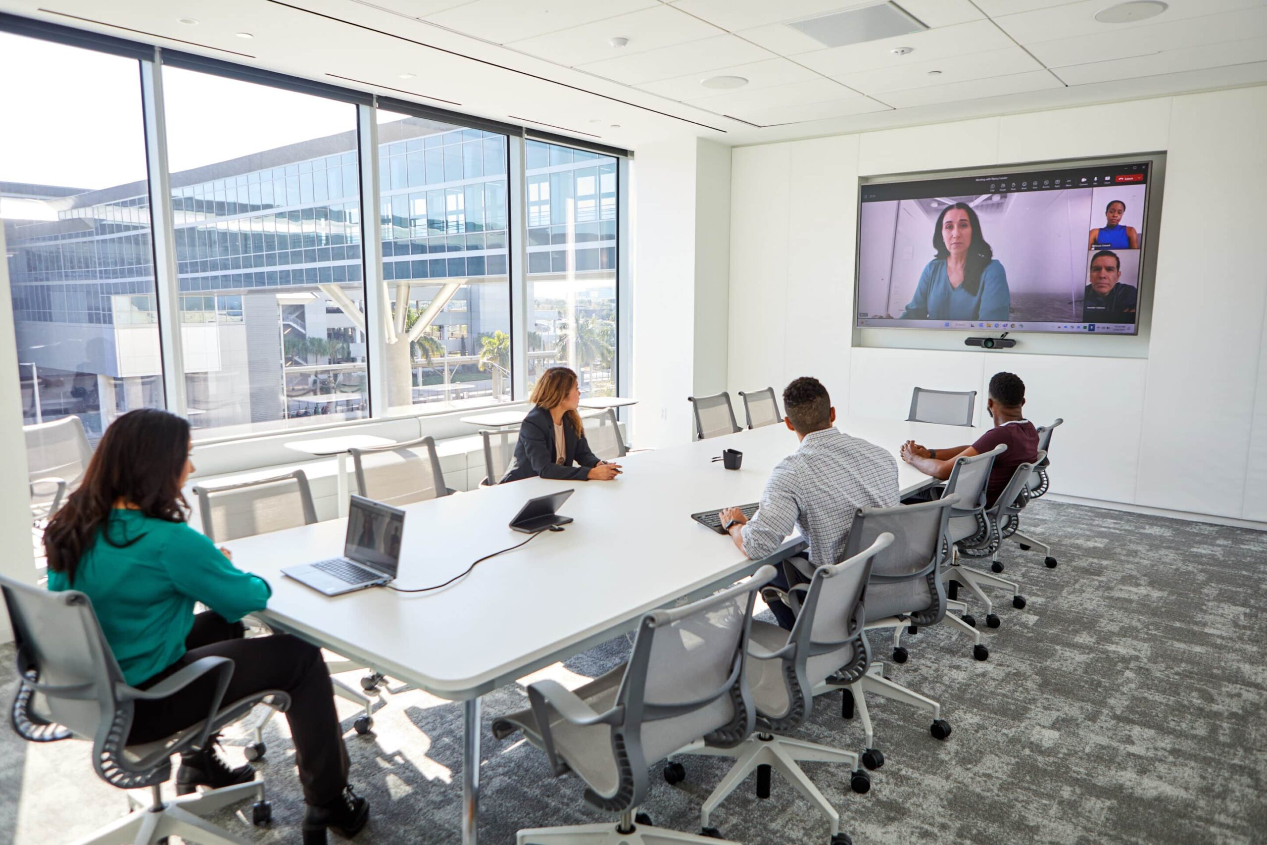 A modern conference room with four people at a table, viewing a video call with three participants; windows show an adjacent building.