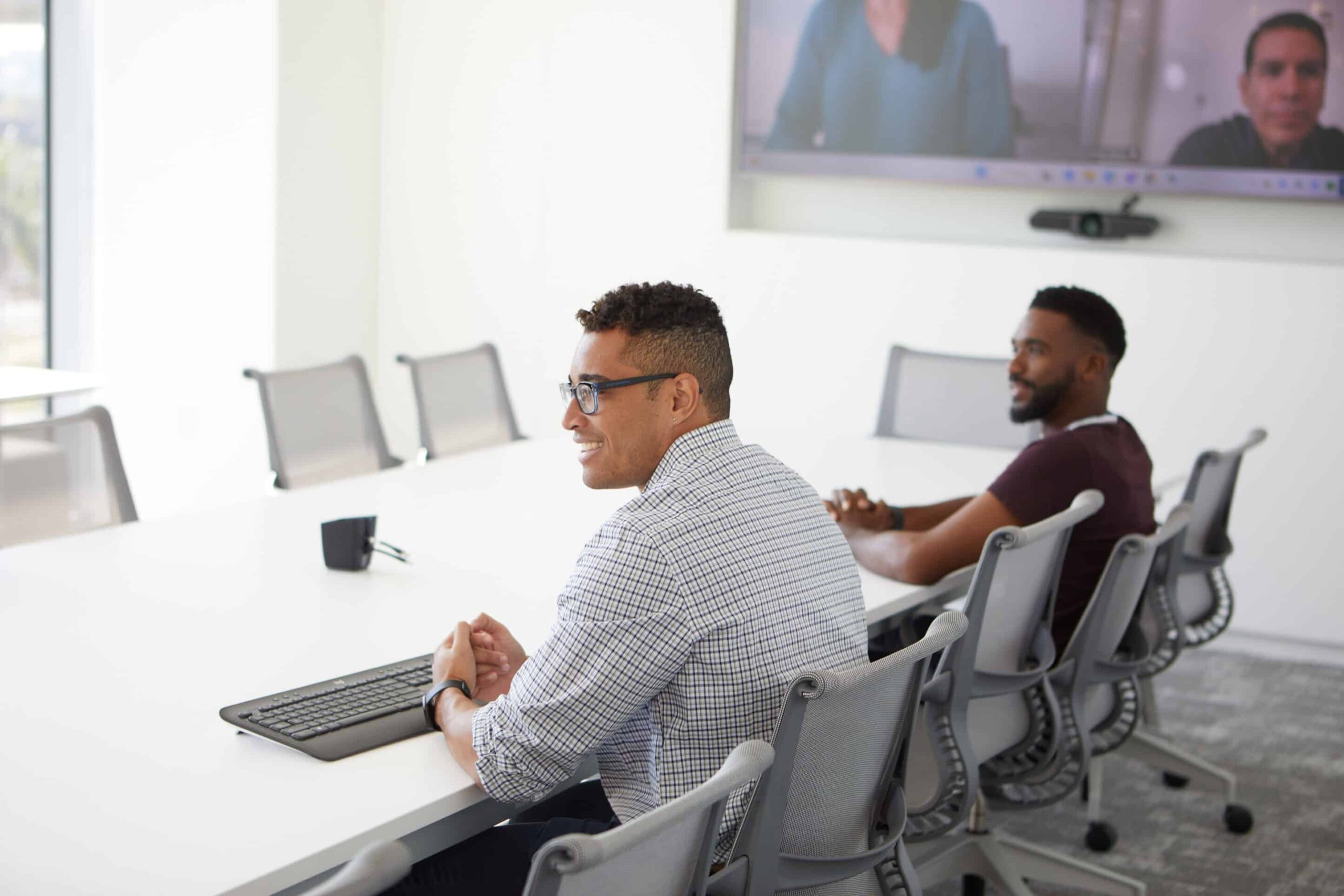 Two people at a conference table, smiling at a video call on screen. Bright, modern room with natural light.