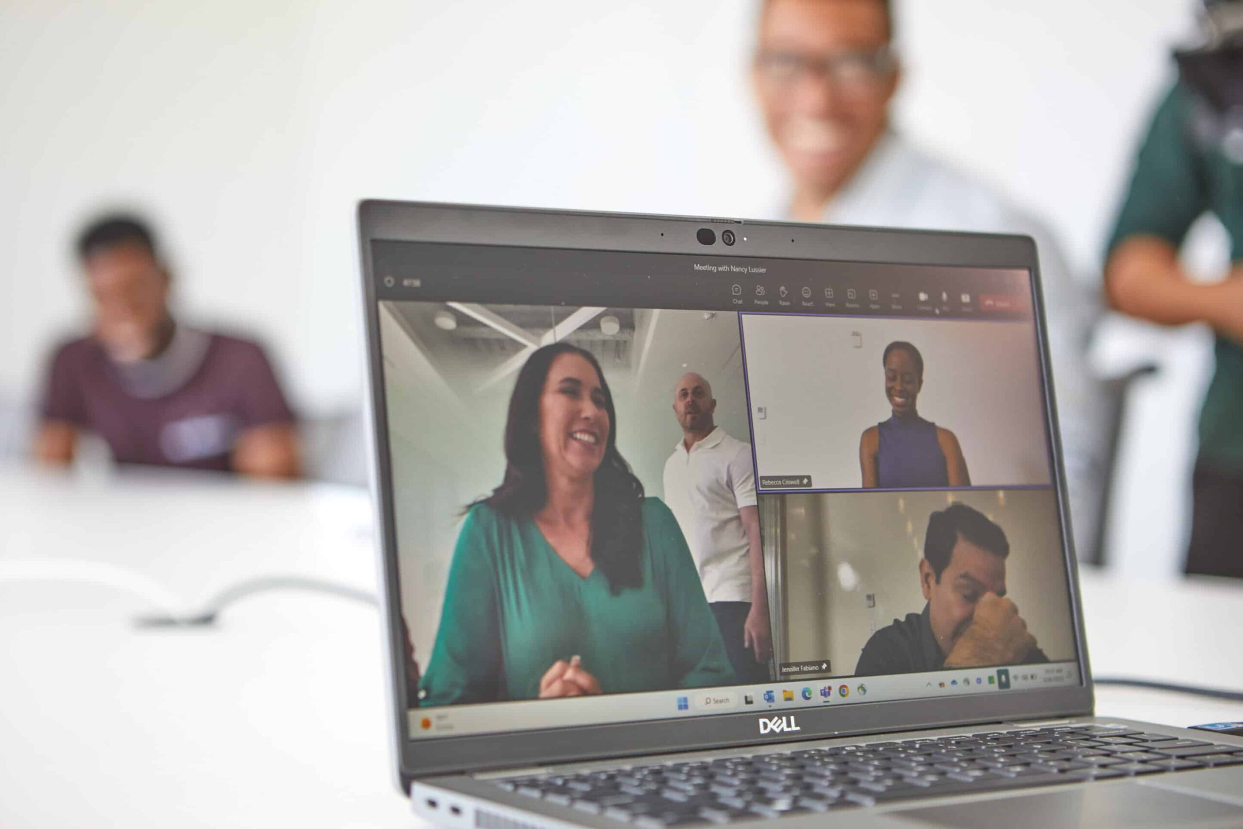 Laptop screen shows a video call with four participants; a woman speaks as others listen. Two blurred figures sit in the background.