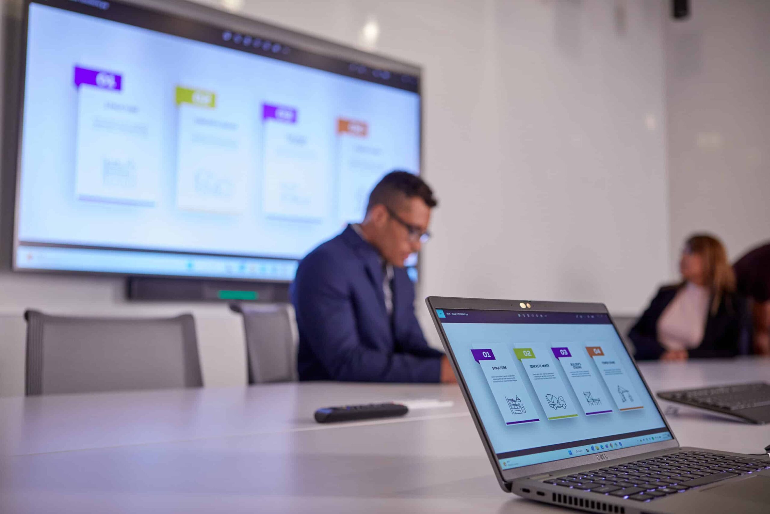 Laptop on table with presentation; person by large screen with unfocused colorful slide; another person partially visible.