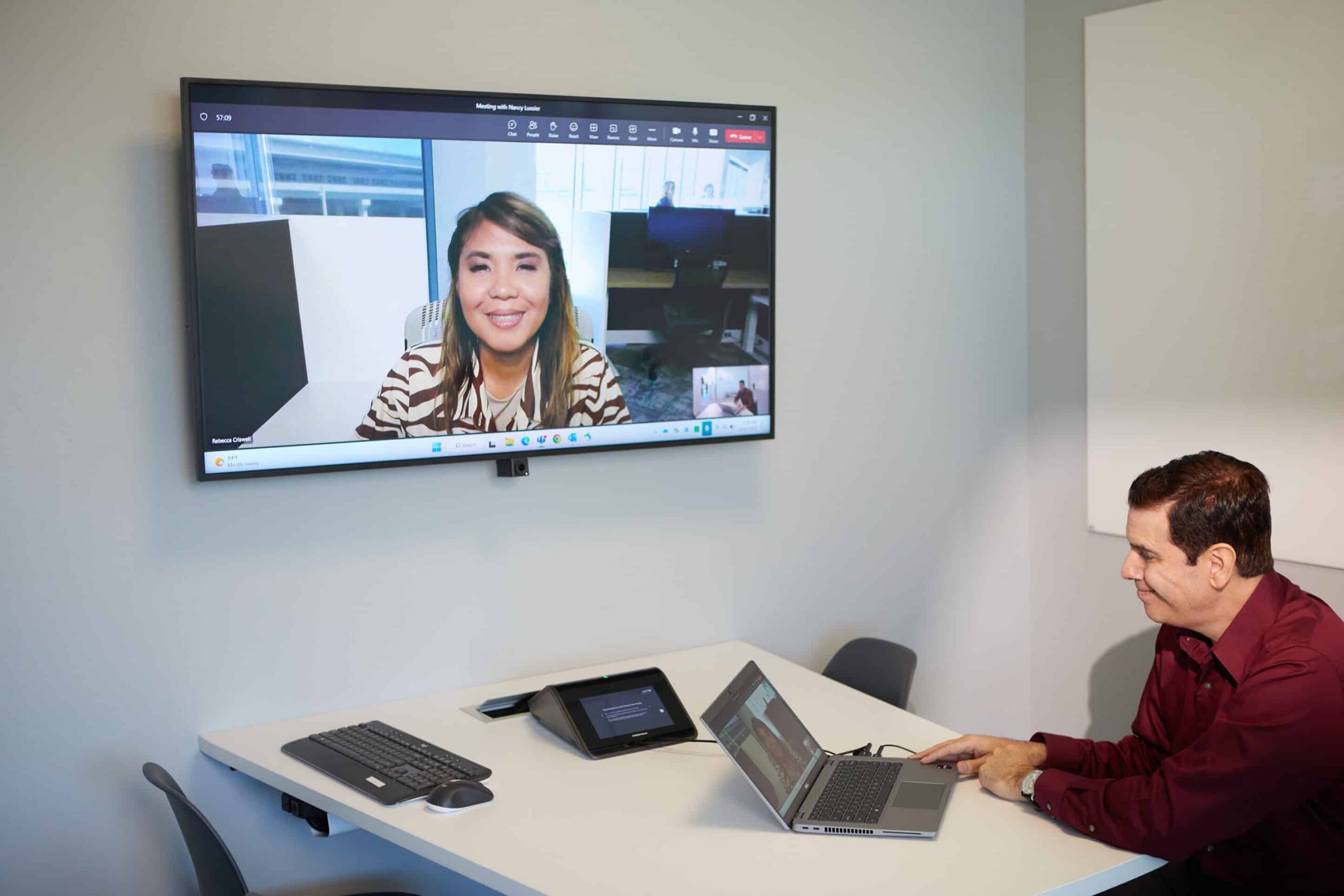 Man in burgundy shirt on video call at conference table; woman smiles on wall screen. Minimalist office setting.