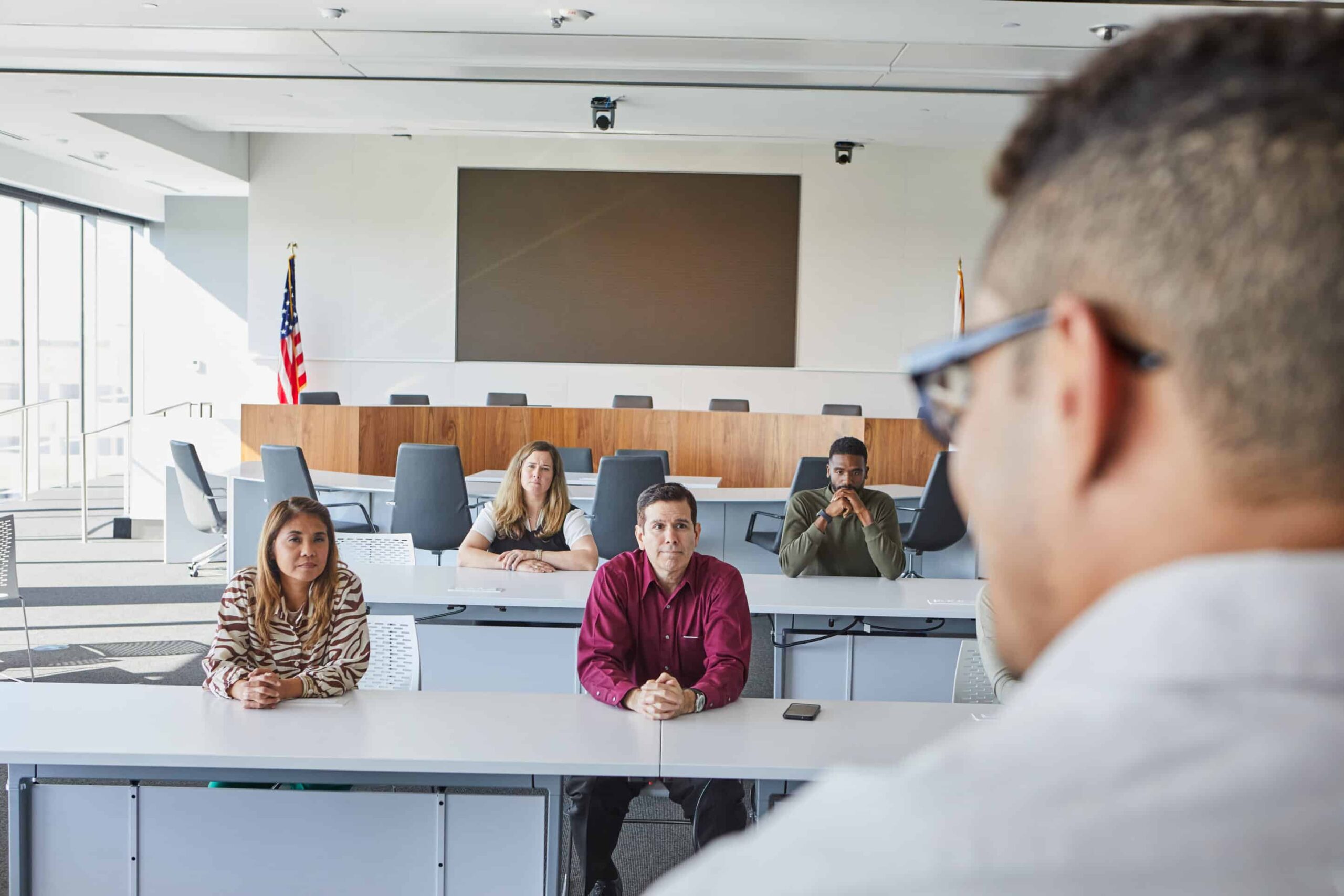 A group sits at desks in a modern conference room, facing a speaker. American flag and large screen visible. Room is well-lit naturally.