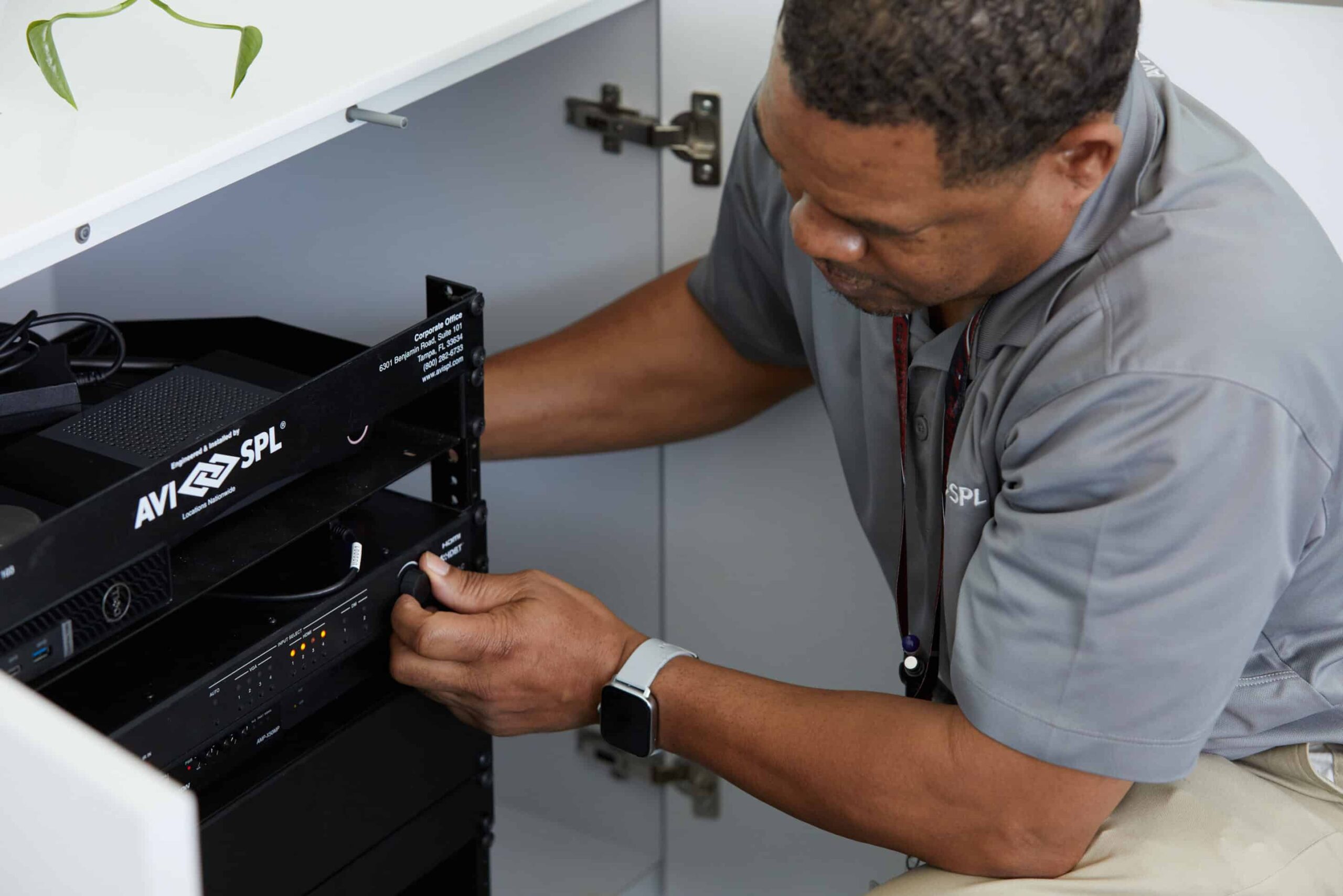 A person in a gray shirt kneels to adjust electronic equipment inside a cabinet, focused on connecting a device, with a plant above.
