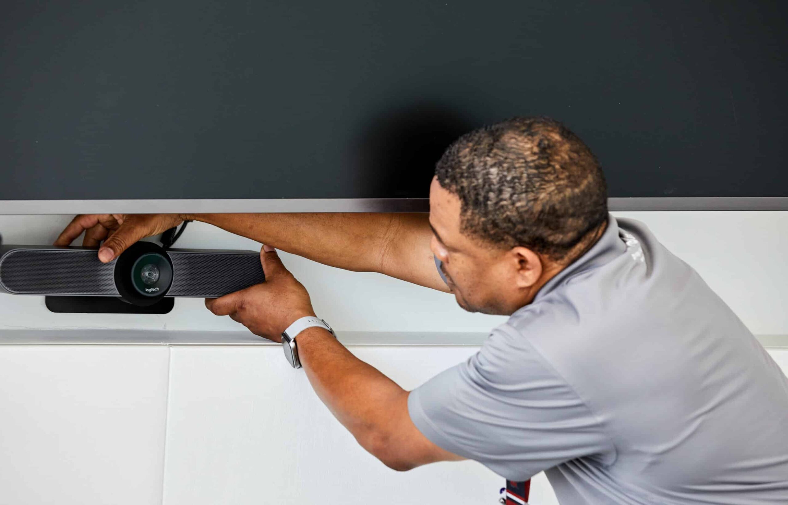 A person in a gray shirt adjusts a video camera under a large screen in a modern, professional room.