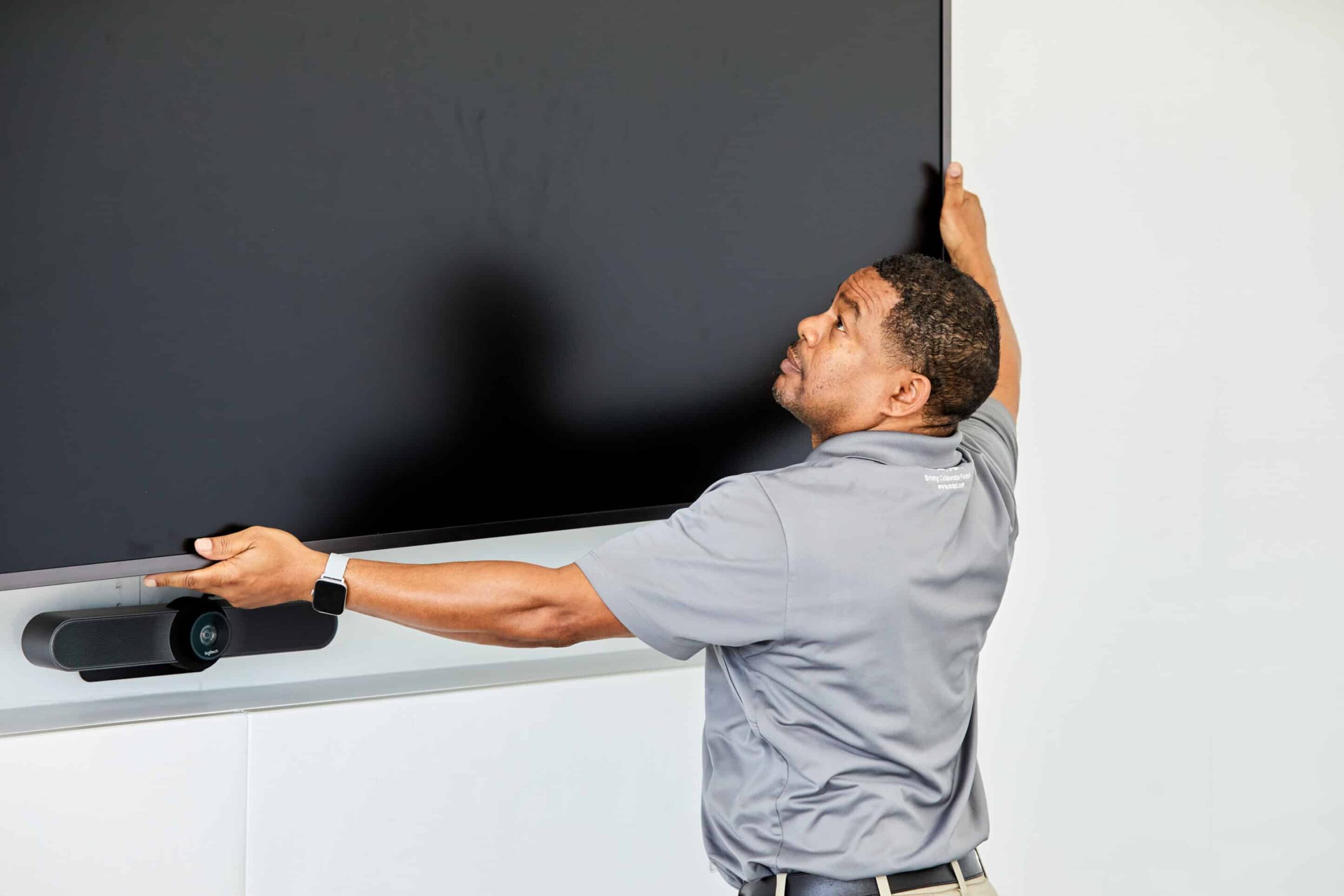 A man in a gray polo mounts a flat-screen TV on a white wall, adjusting it with both hands; a small device is below the screen.