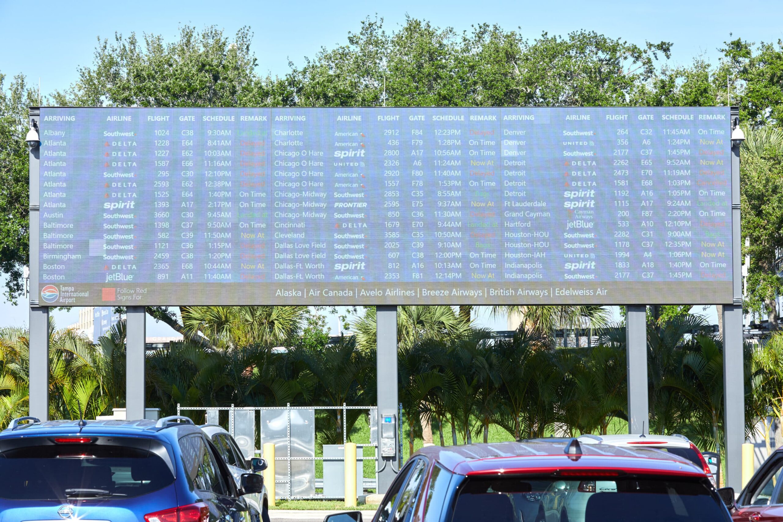 Airport arrivals board shows flights with airline, origin, flight number, times, gate, baggage info; trees and cars below.