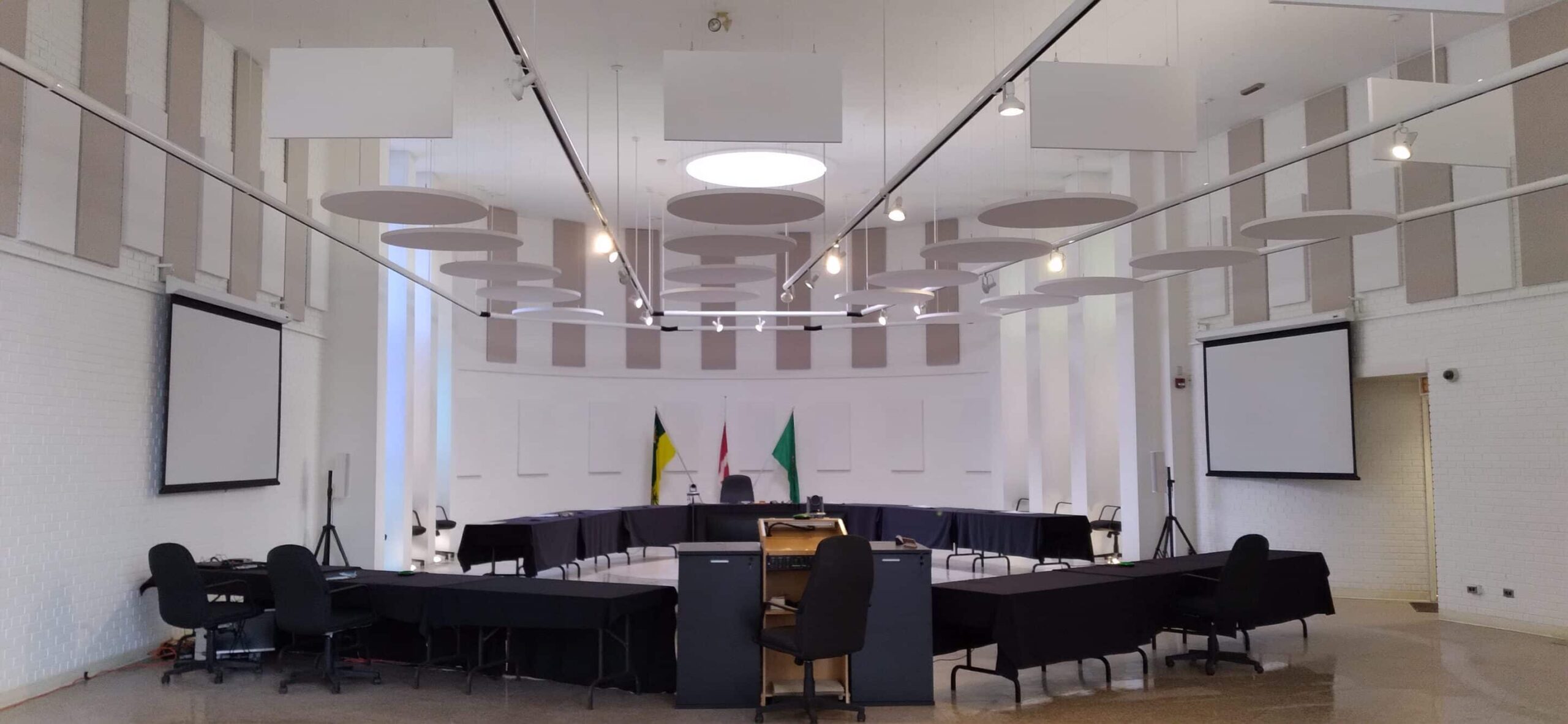 Modern conference room with a large table, black chairs, flags, circular ceiling panels, lights, and two blank side projector screens.