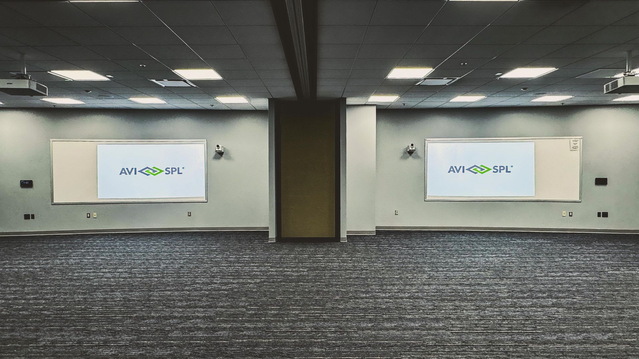 Spacious modern conference room with gray decor; two AVI SPL screens, ceiling lights, no furniture visible.