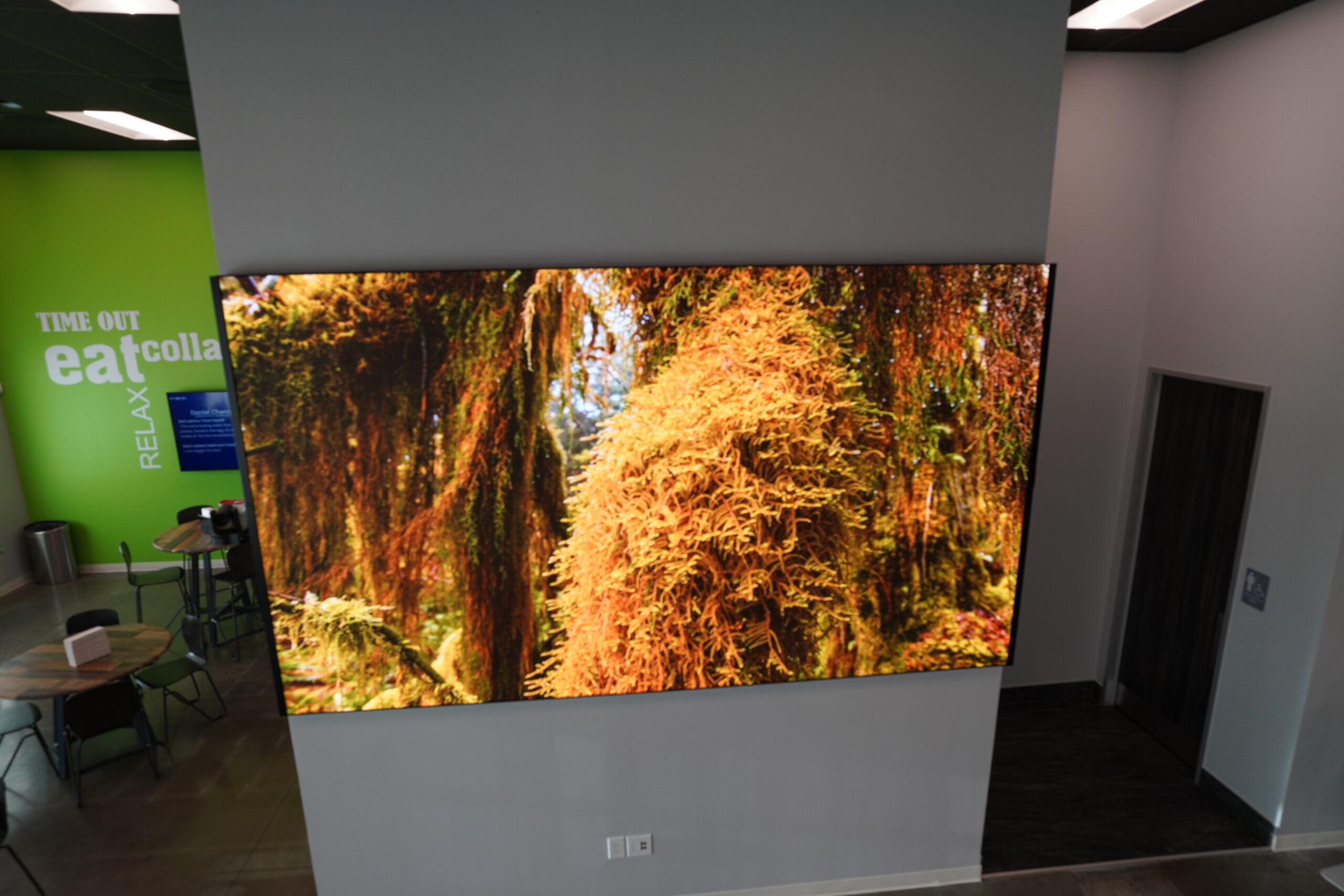 A large digital screen displays a vibrant image of lush, green and orange foliage in a modern room. Walls are gray with tables and chairs nearby. Text on a green wall reads TIME OUT eat relax.