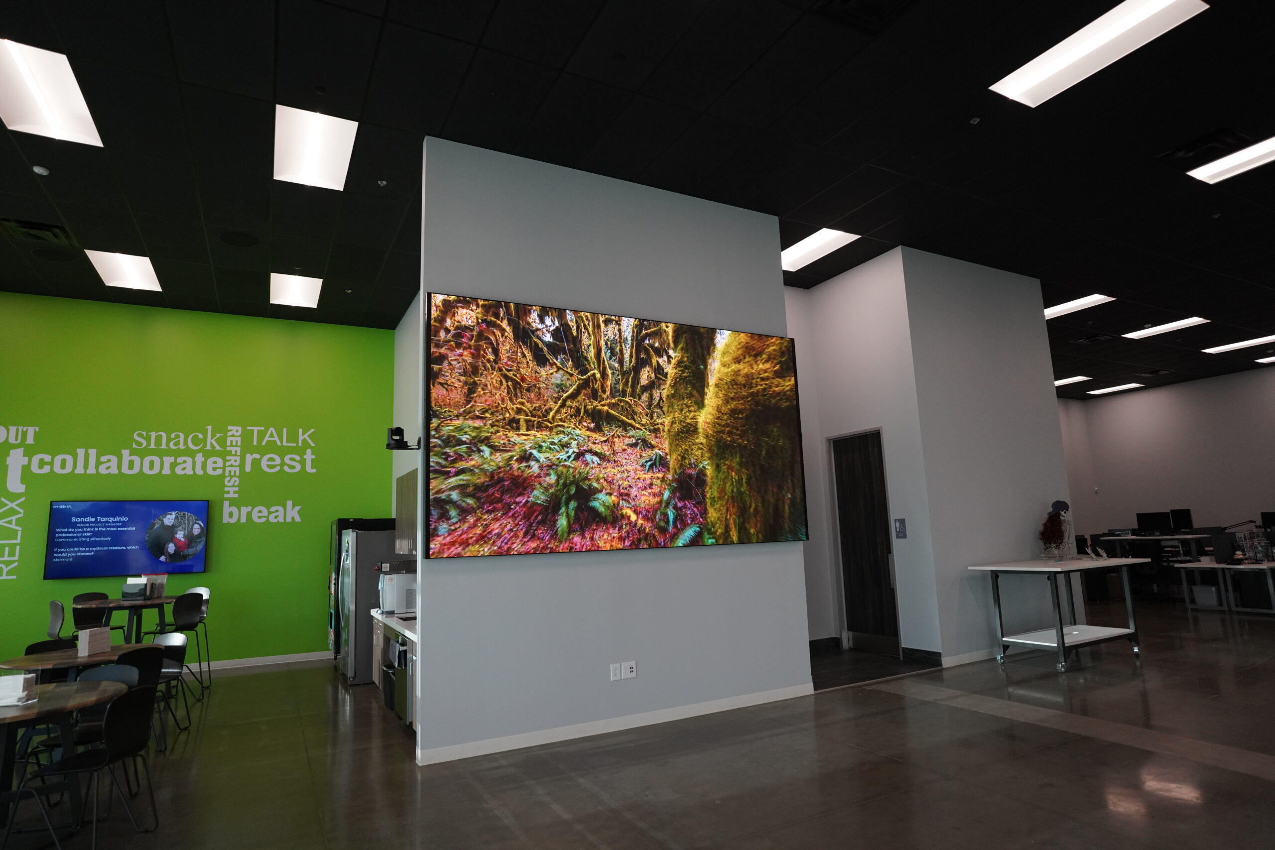 Modern office with a digital nature scene, green/white walls with collaborate and snack, tables, chairs, and person at desk.