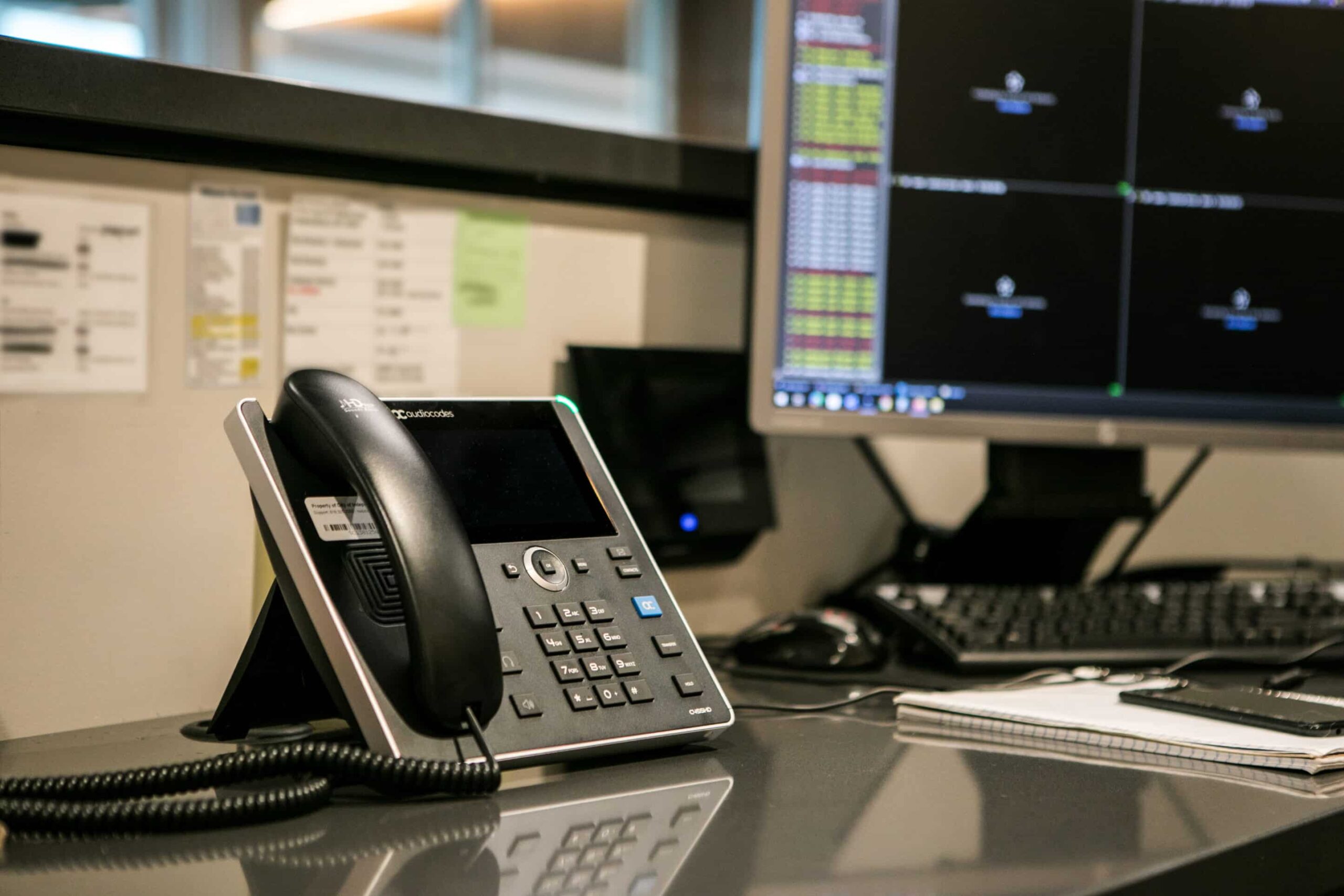 Close-up of an office desk with a black phone, monitor showing data charts, papers, notebook, and keyboard in a typical setup.