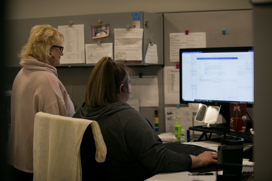 Two people in an office: one works at a cluttered desk with a computer and headset, the other stands nearby.