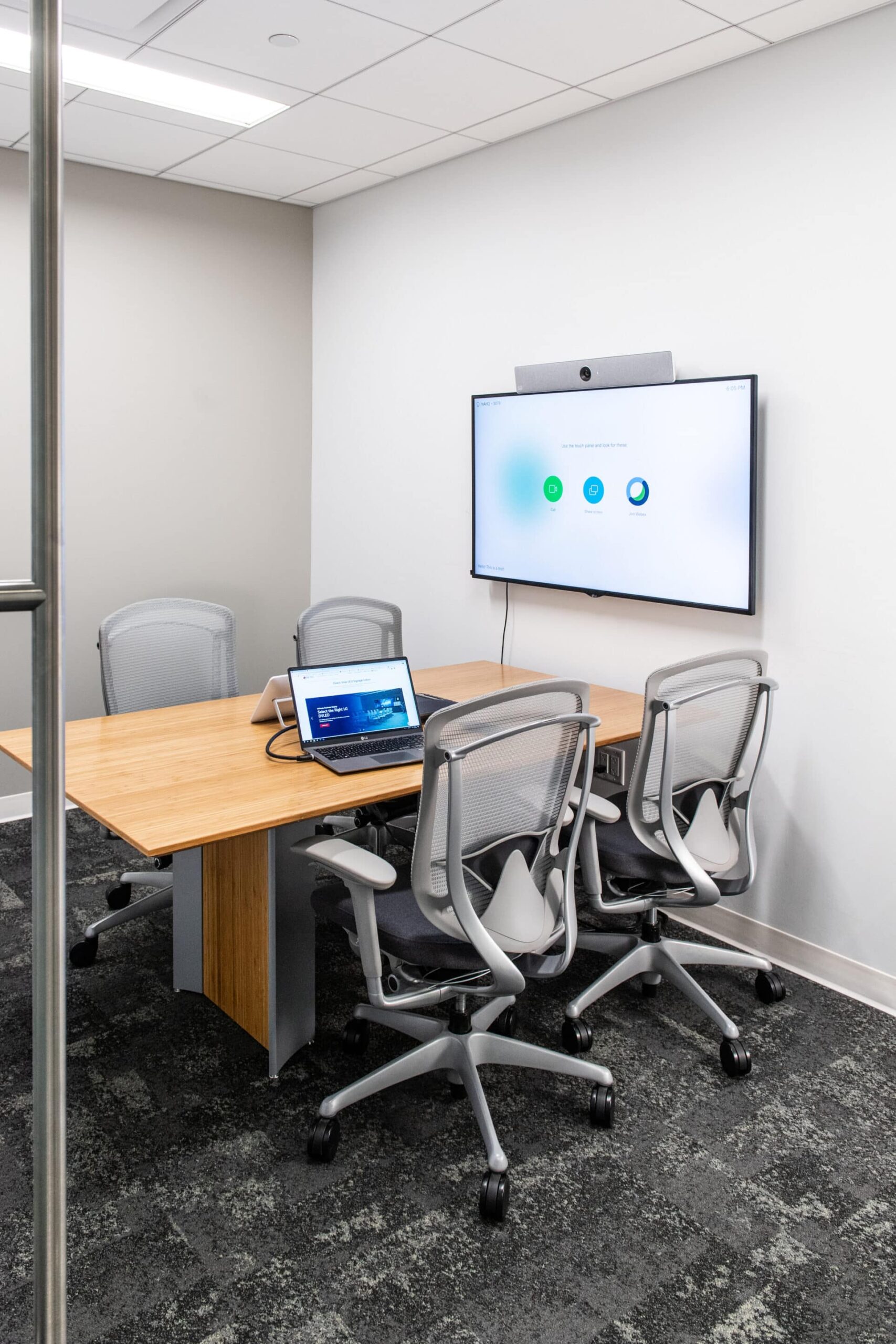 Modern conference room: wooden table, four gray chairs, screen with video call, laptop with slides, gray carpet, white walls.