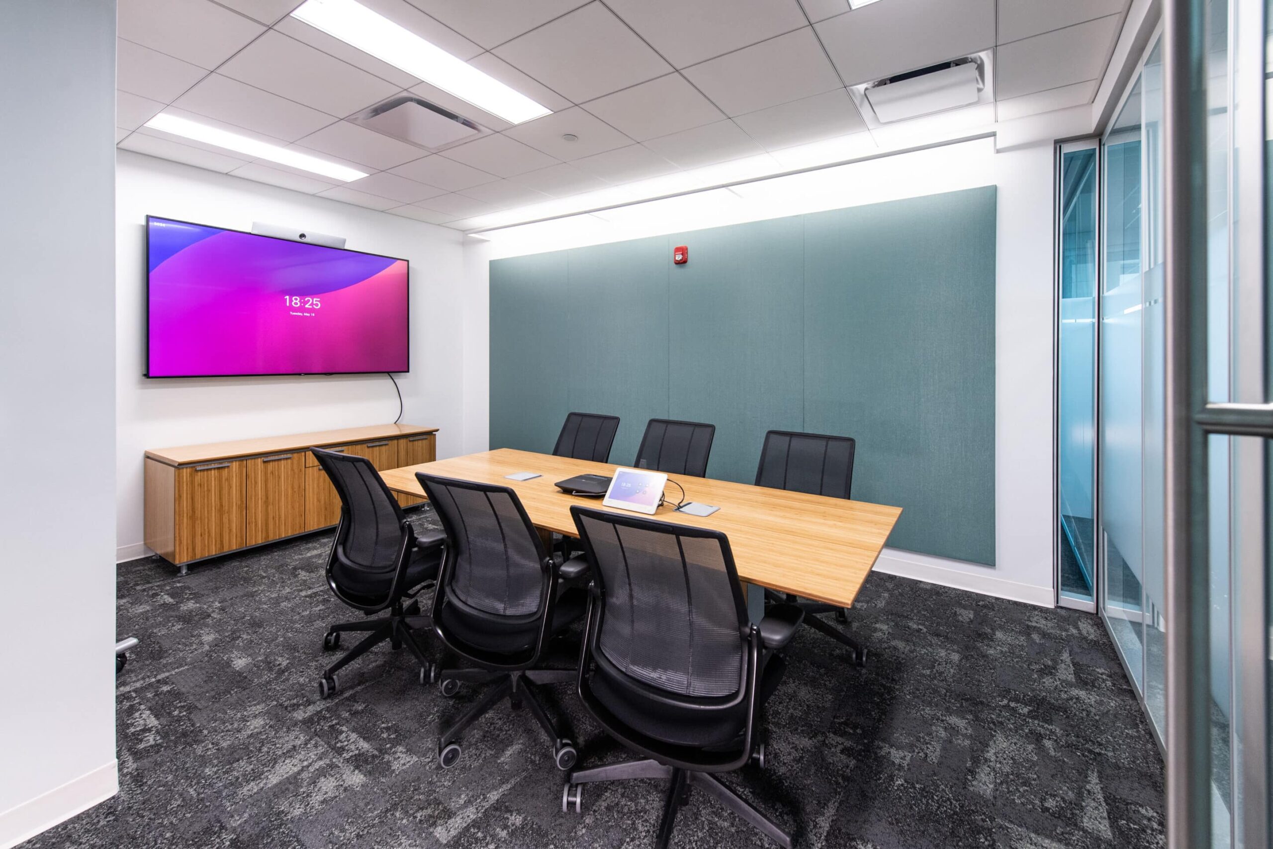 Modern conference room: wooden table, 5 black chairs, large screen showing 18:25. Grey carpet, glass panel wall.