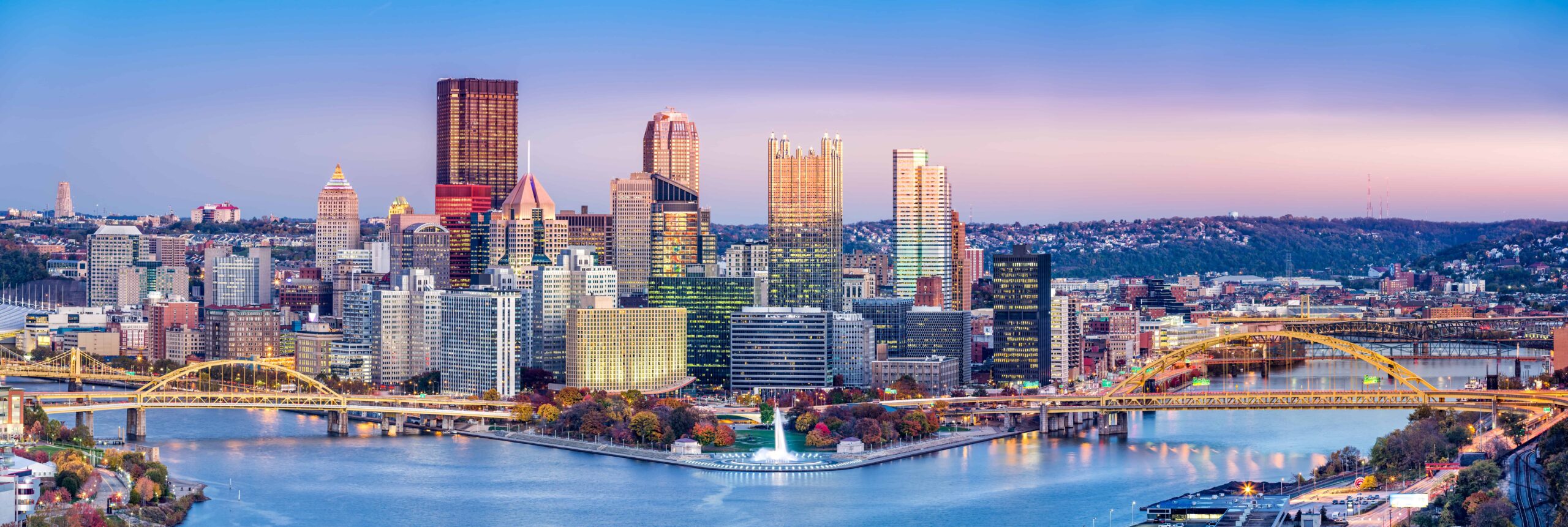 City skyline at sunset with modern high-rises, bridges, a river, and autumn trees; clear sky with pastel hues on the horizon.