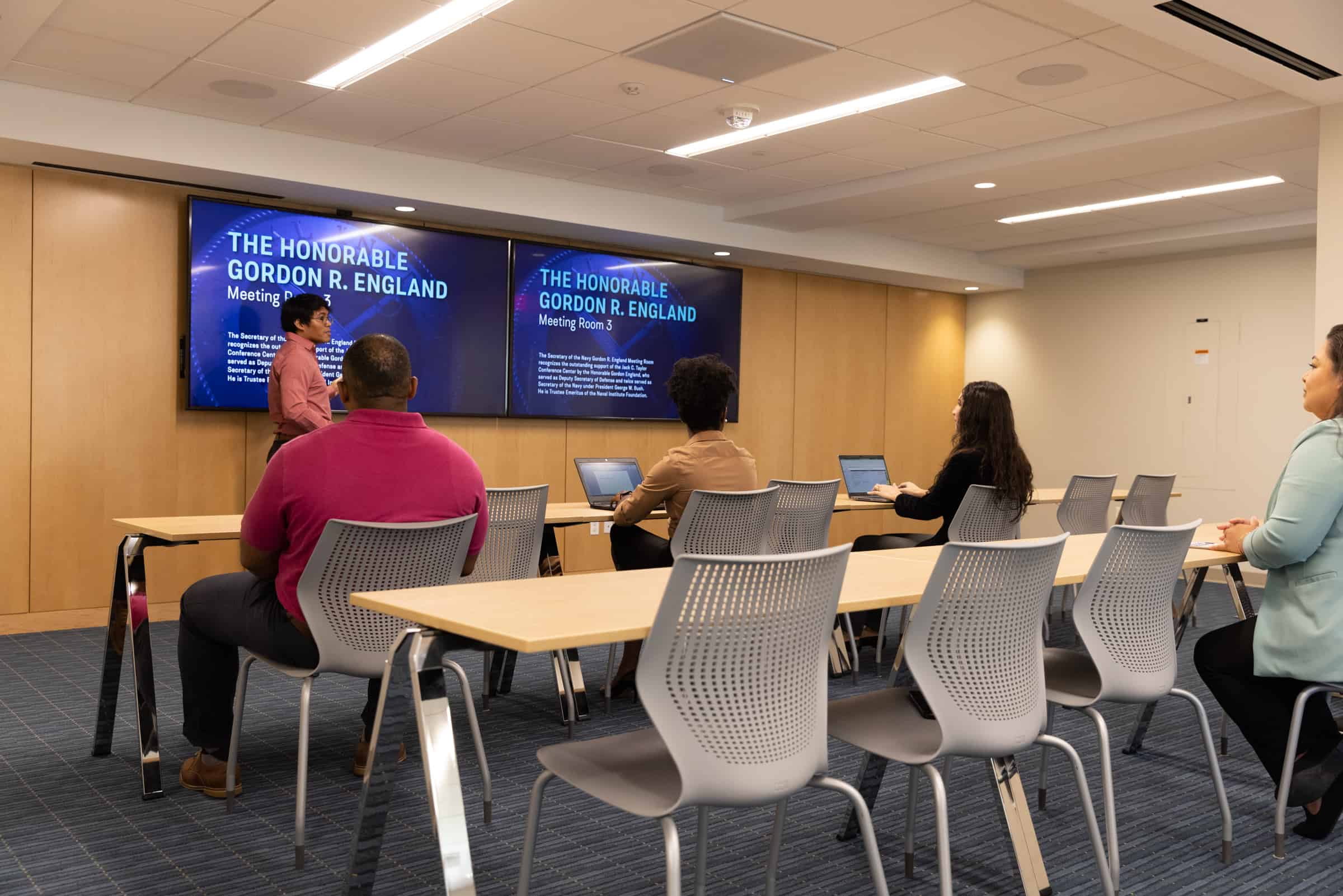 A person presents in a modern conference room with attendees at desks. Two large screens display The Honorable Gordon R. England.