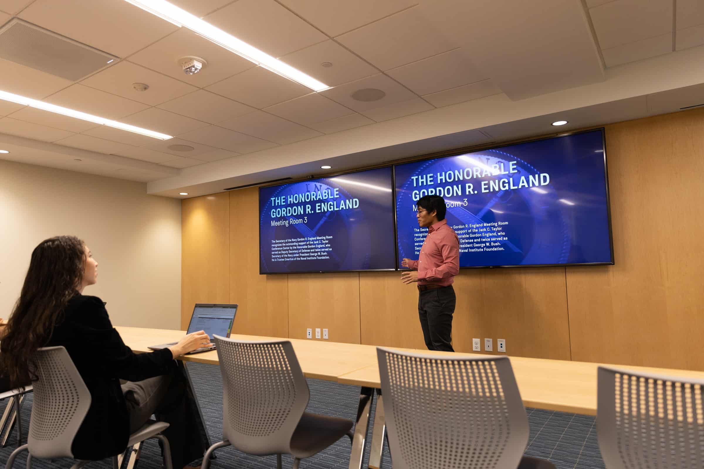 A speaker in a pink shirt presents The Honorable Gordon R. England as a woman with a laptop listens in a conference room.