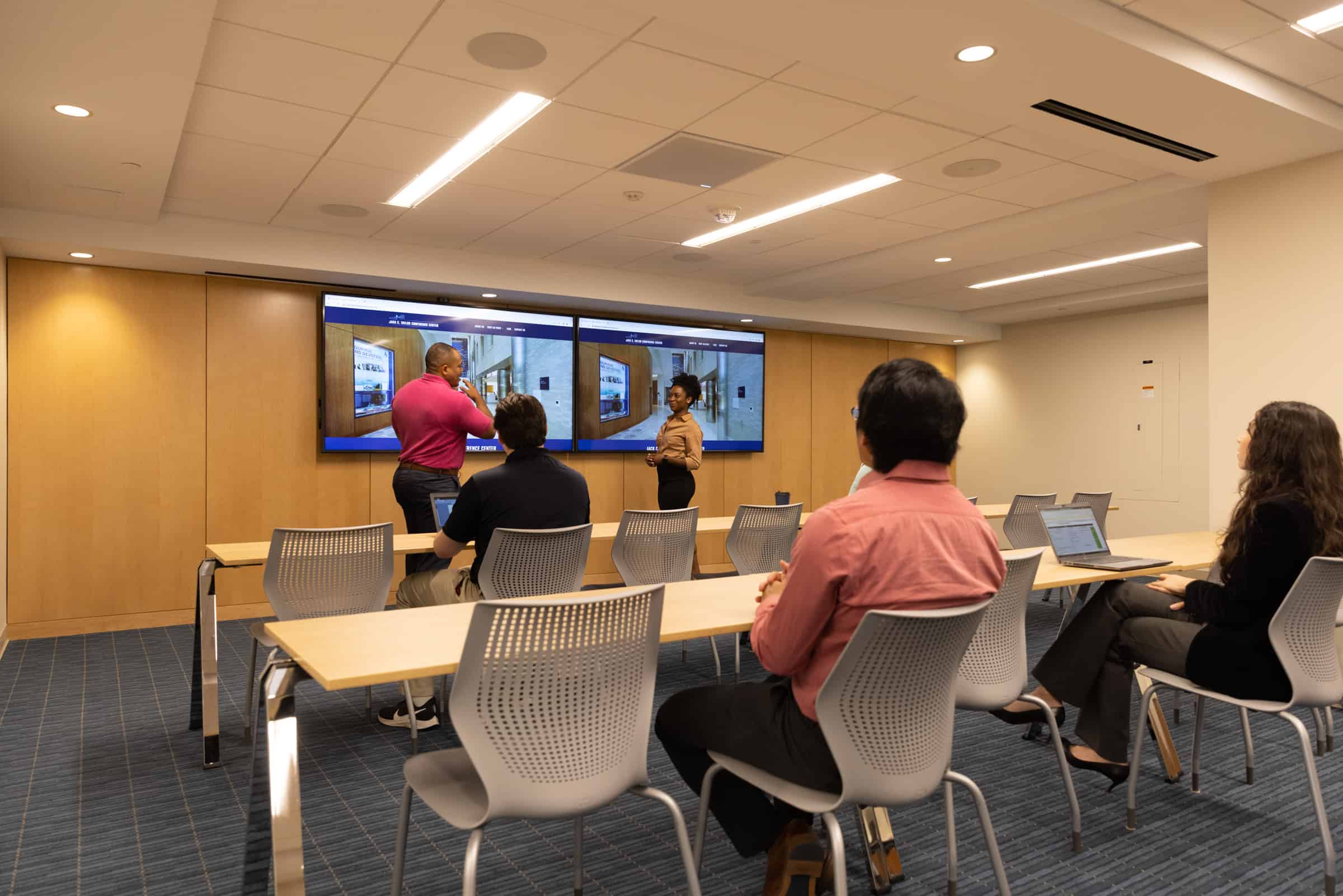 Office group near screen with images; two leading discussion. Modern room, blue carpet, beige walls.