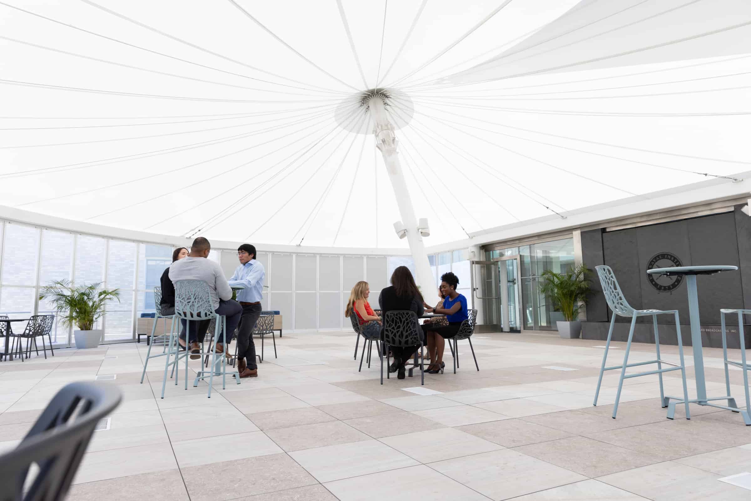 People gather in a bright, open space with a white canopy ceiling, modern furniture, and plants, chatting around tables.