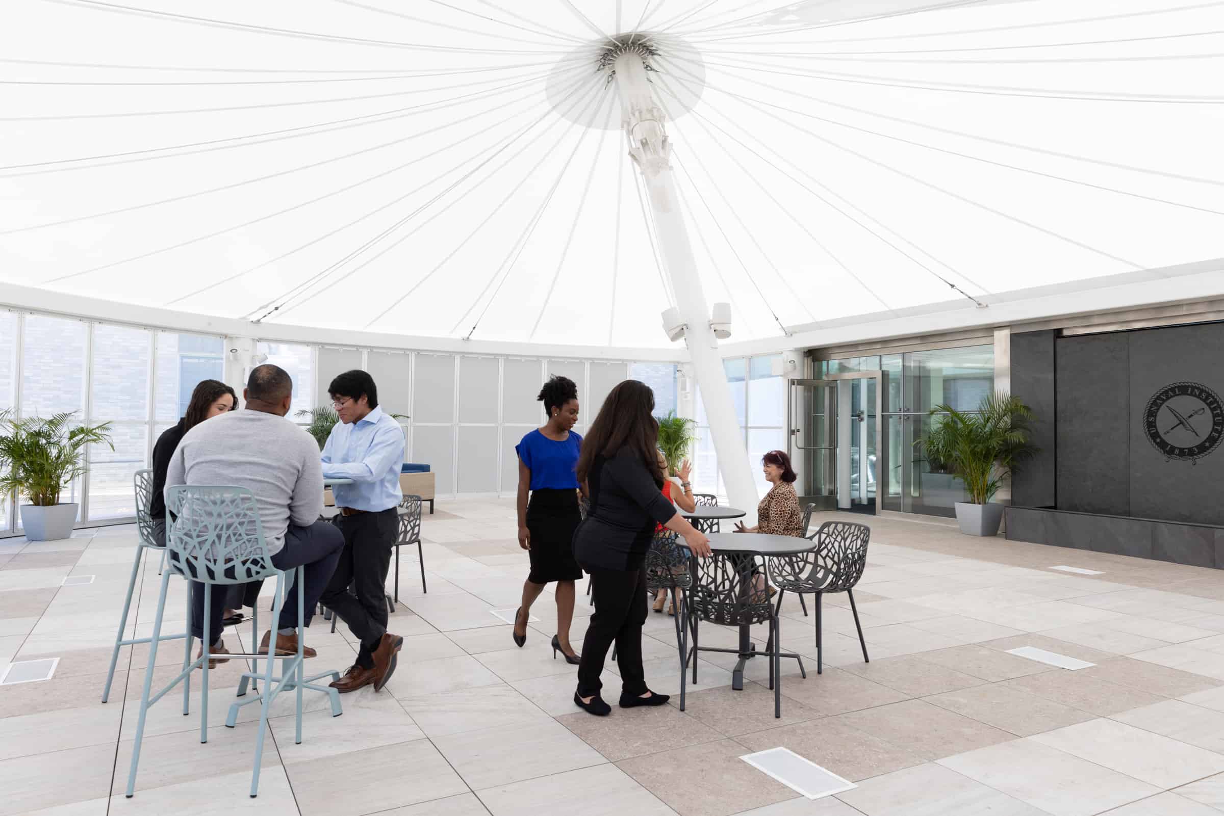 People gather in an open-air lounge, chatting at tables. The space has metal chairs, decorative plants, and a clear ceiling structure.