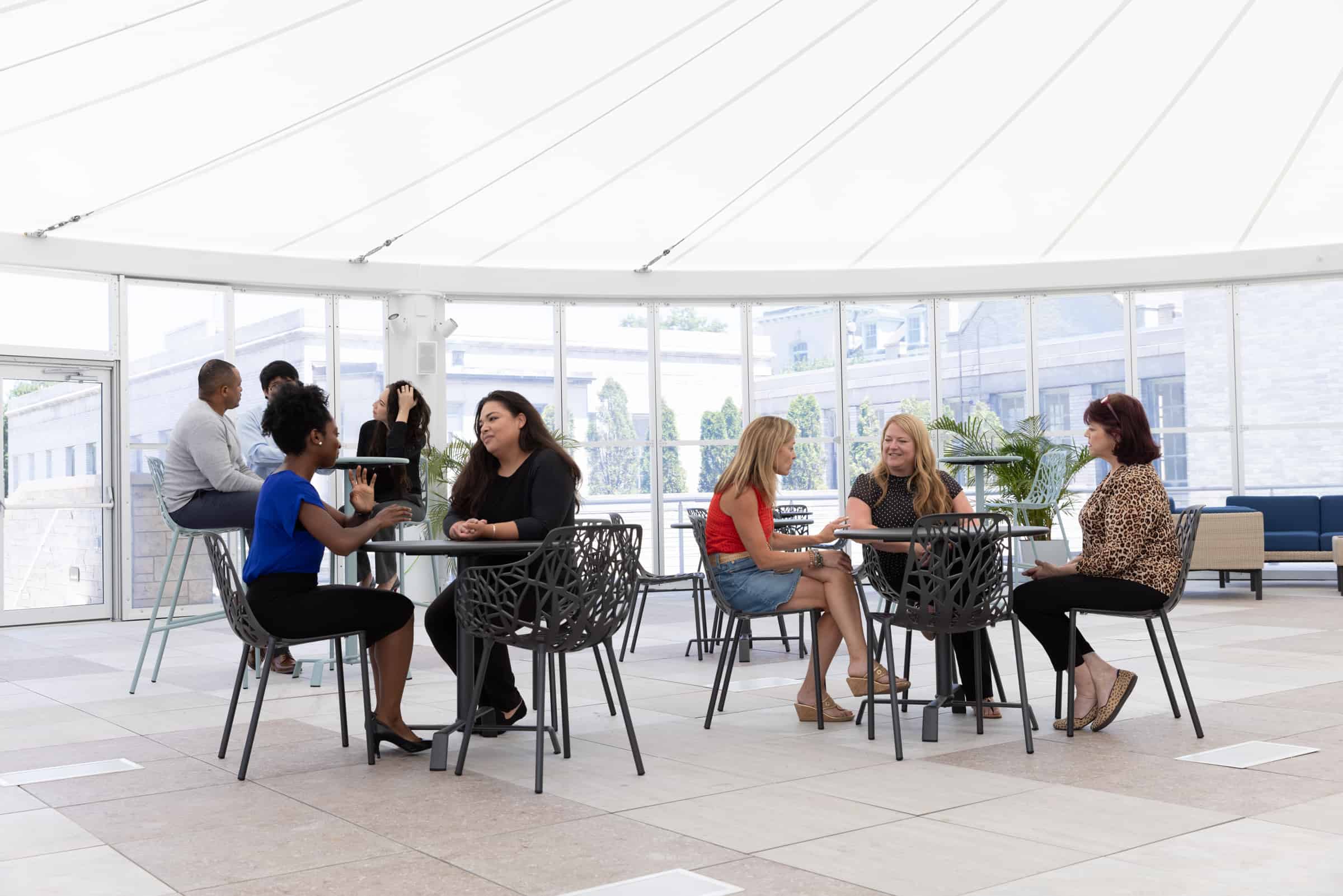 People chat at tables in a bright, modern rooftop terrace with large windows and a unique white canopy ceiling.