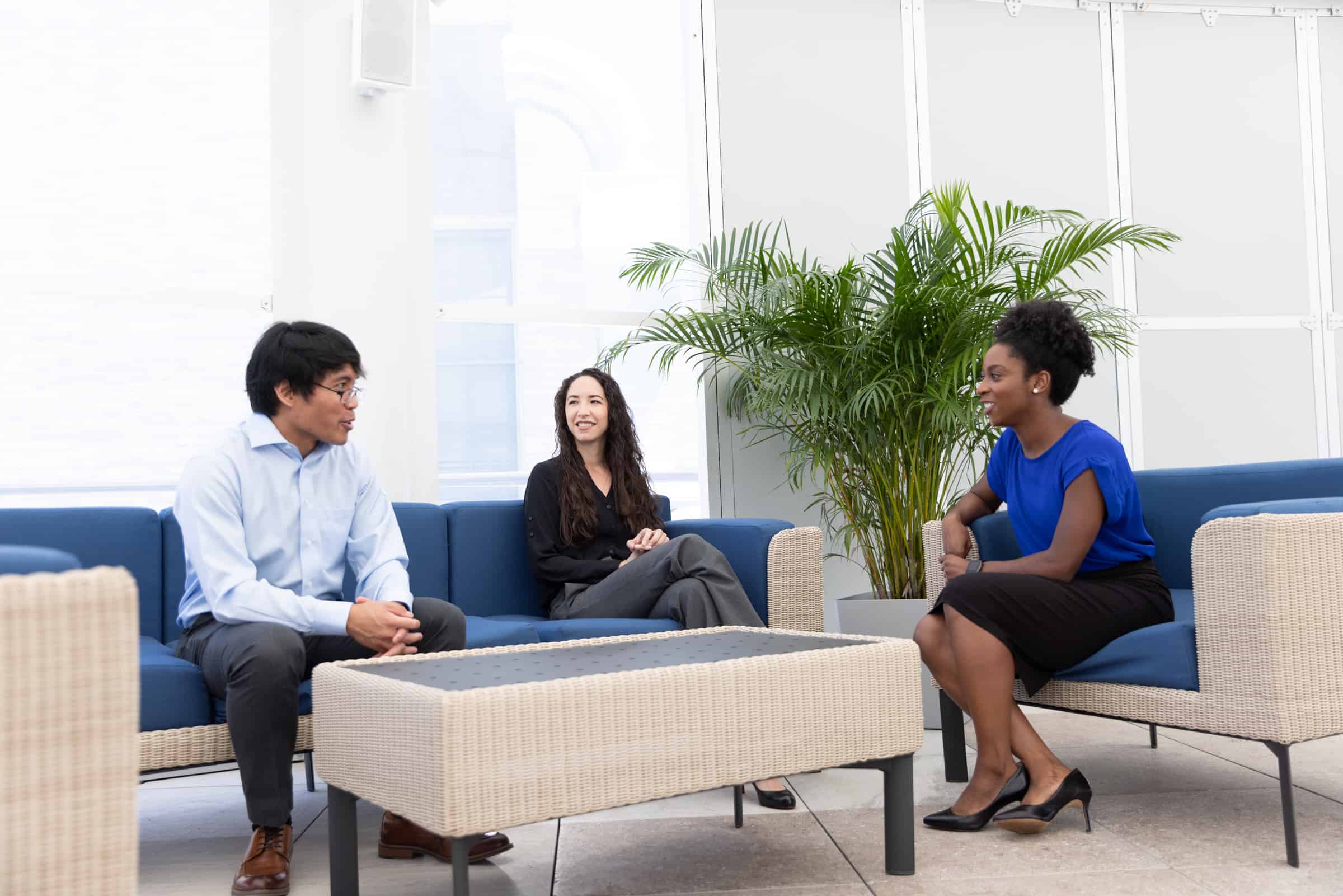 Three people chat in a modern lounge with blue couches and a green plant, dressed in business casual.