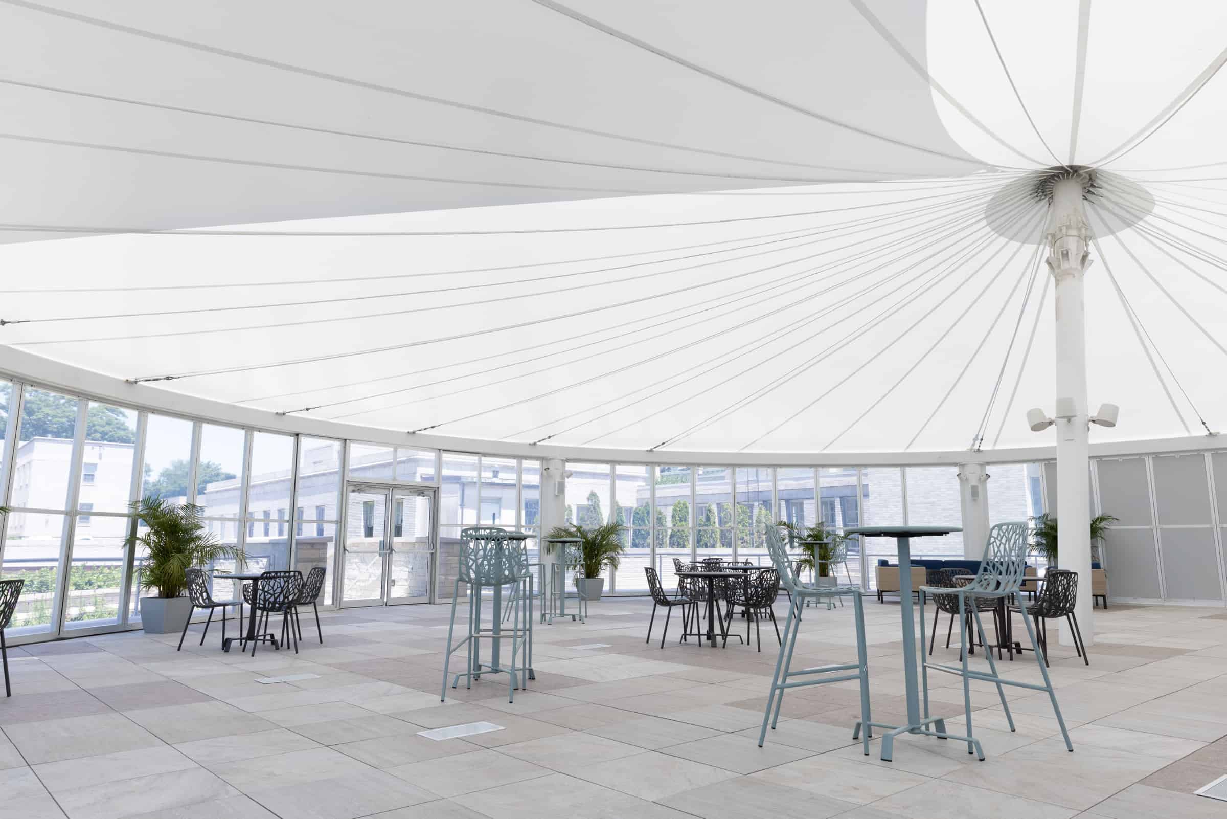 A spacious indoor area with a tent-like ceiling, glass walls, tables, chairs, plants, tiled floor; filled with natural light.
