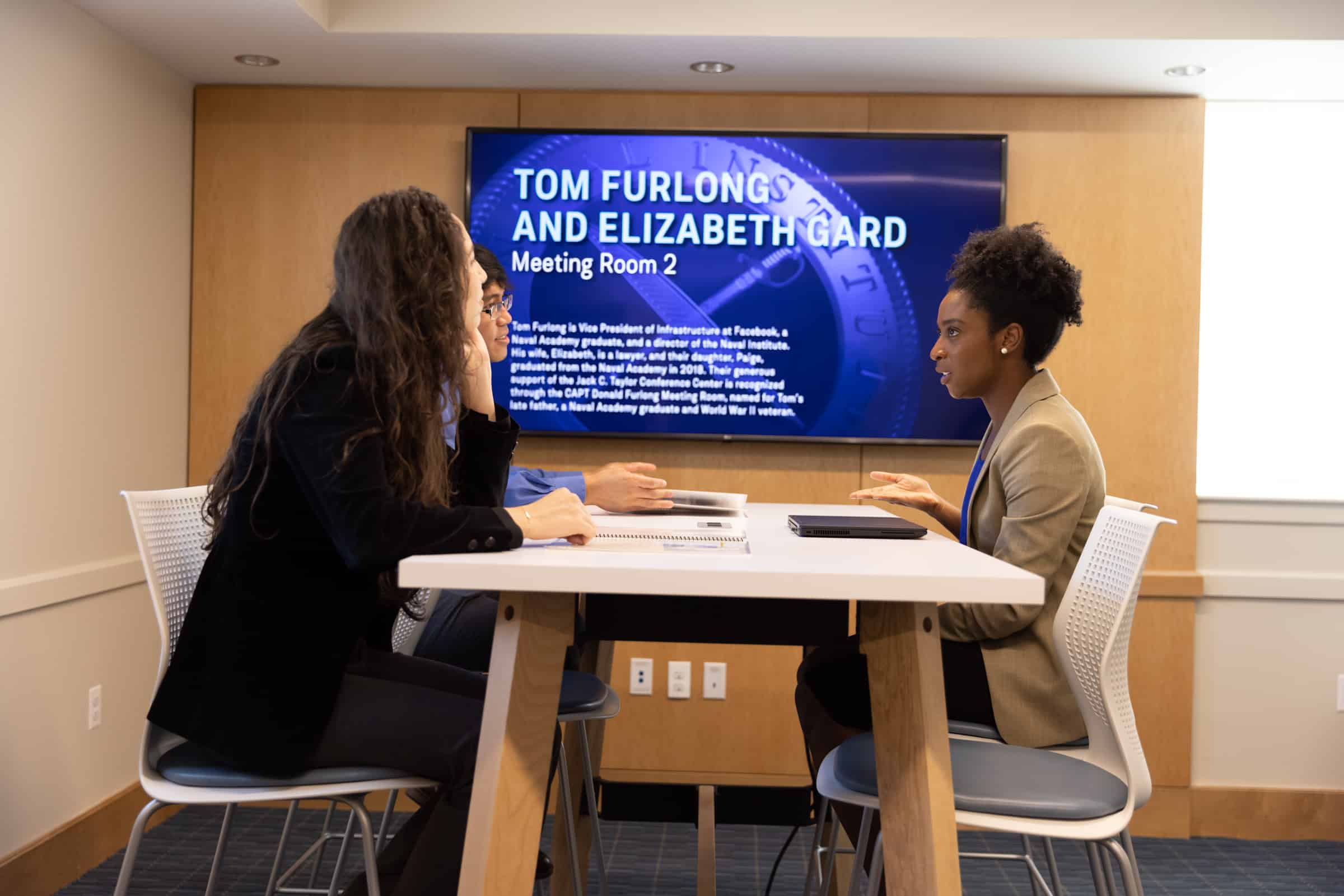 Three people in Tom Furlong & Elizabeth Gard Meeting Room 2 with papers and laptops are engaged in a discussion.