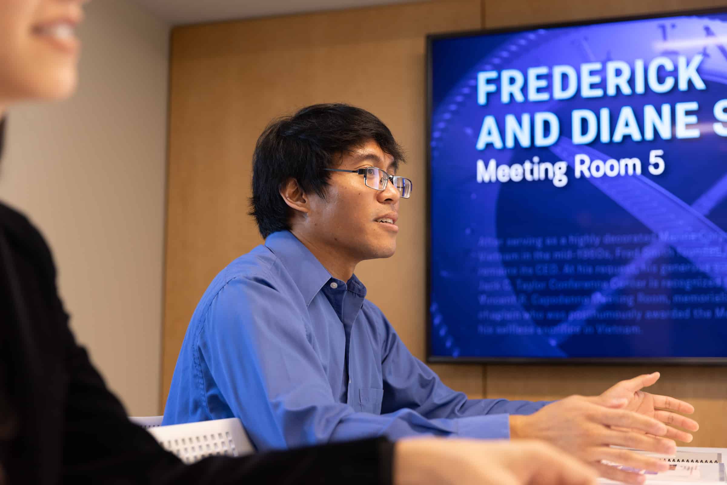 Blue-shirted person speaks at a table in Frederick and Diane S. Meeting Room 5; another figure is partially visible.