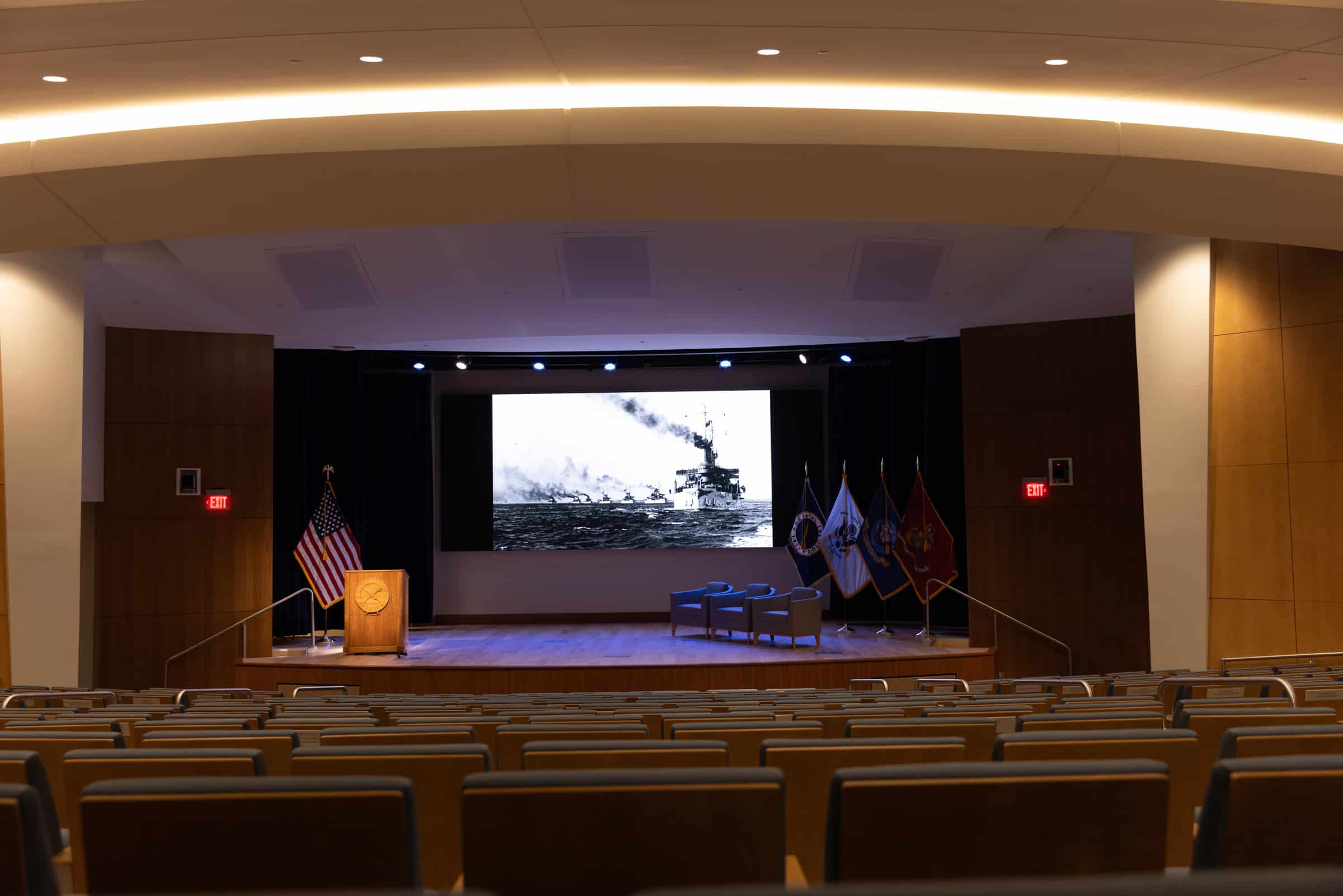 Modern auditorium with empty seats facing a stage; screen shows ship, flags, podium & two chairs present.