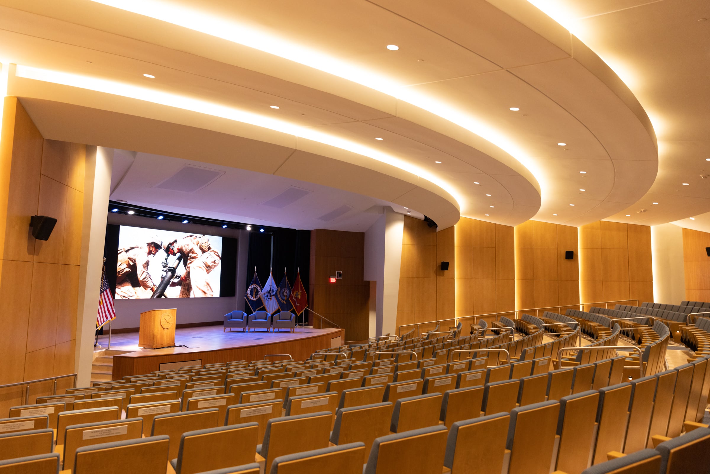Empty auditorium with rows of wooden chairs, a stage with podium, flags, large screen showing people; modern curved lighting ceiling.