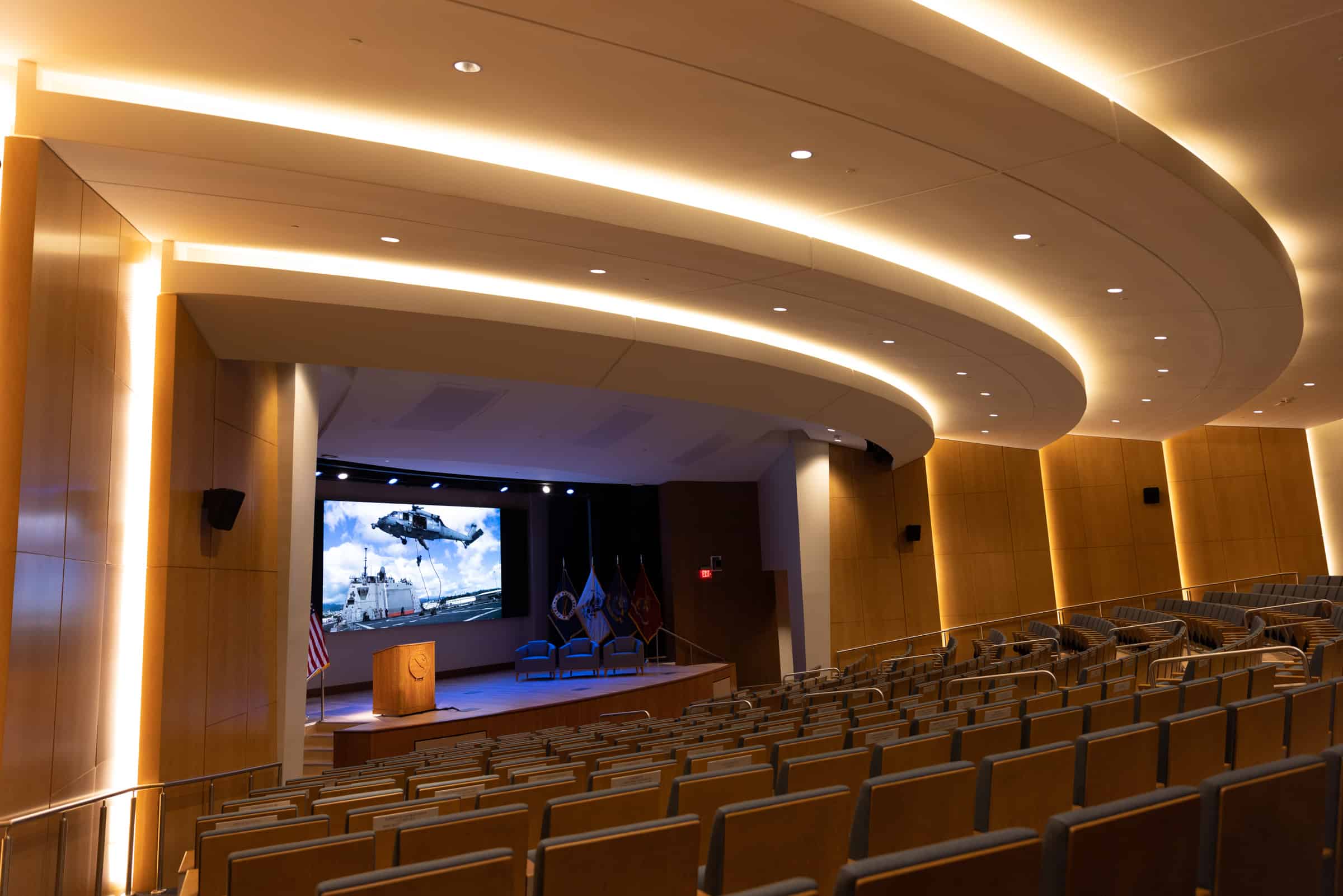 Modern auditorium with empty seats, curved paneling, recessed lights; stage shows a helicopter-ship image, podium flags.