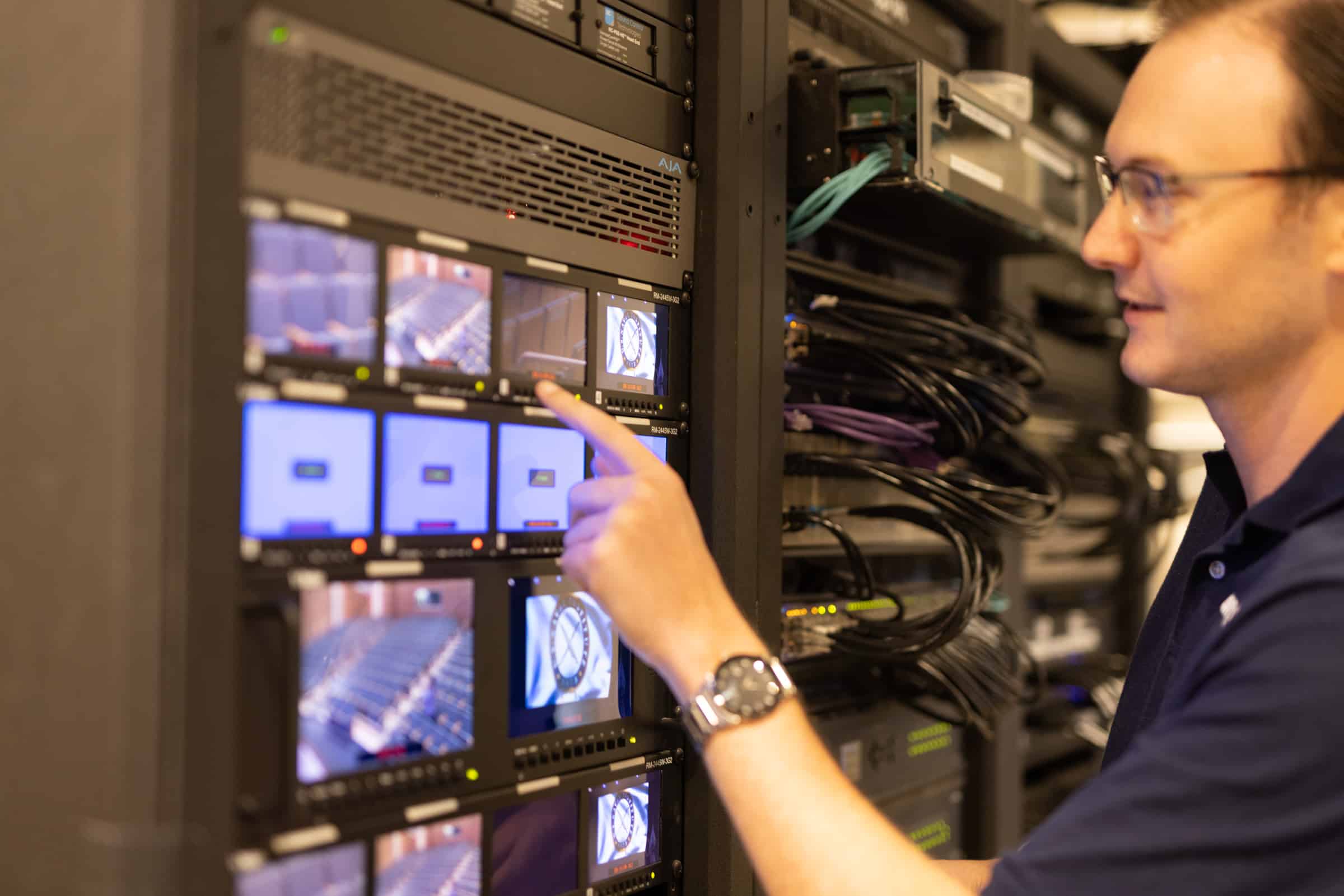 A person in a blue shirt interacts with a server rack full of screens and equipment, displaying technical images and data.