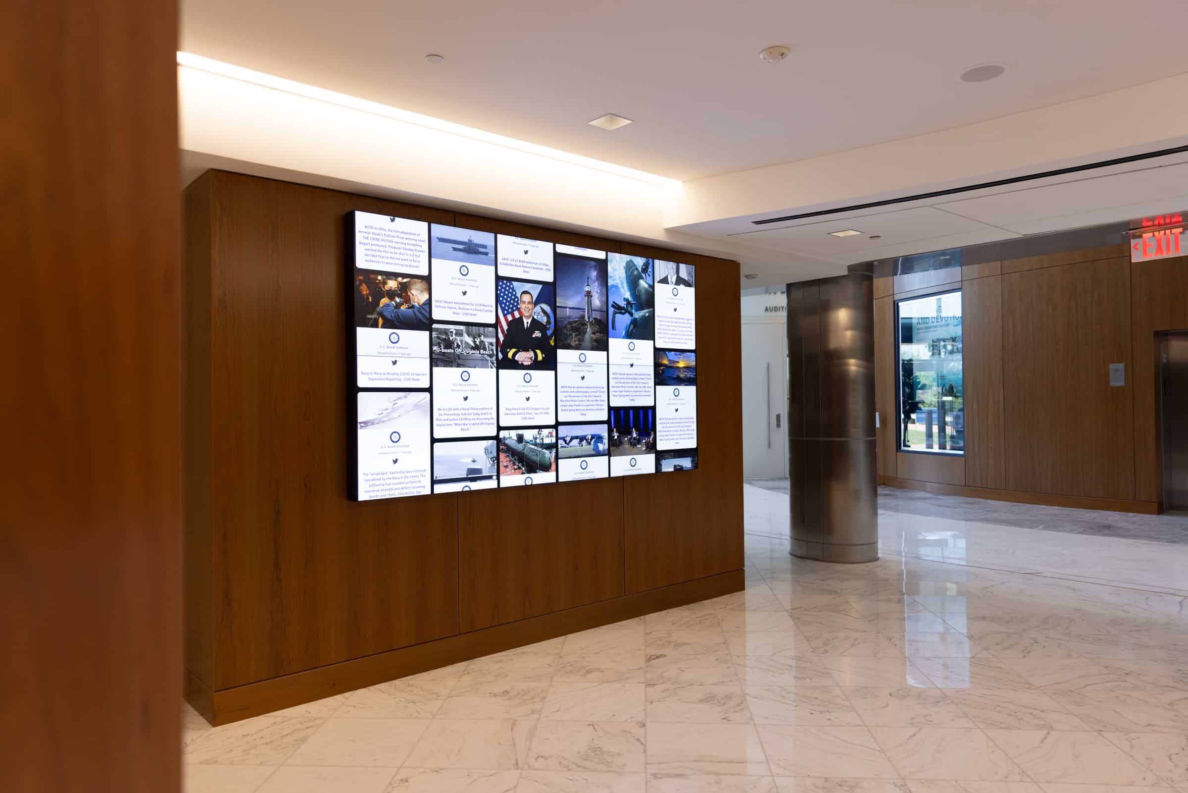 Modern lobby with digital display on wooden wall for news/social feeds, white marble floor, wood accents, glass doors.