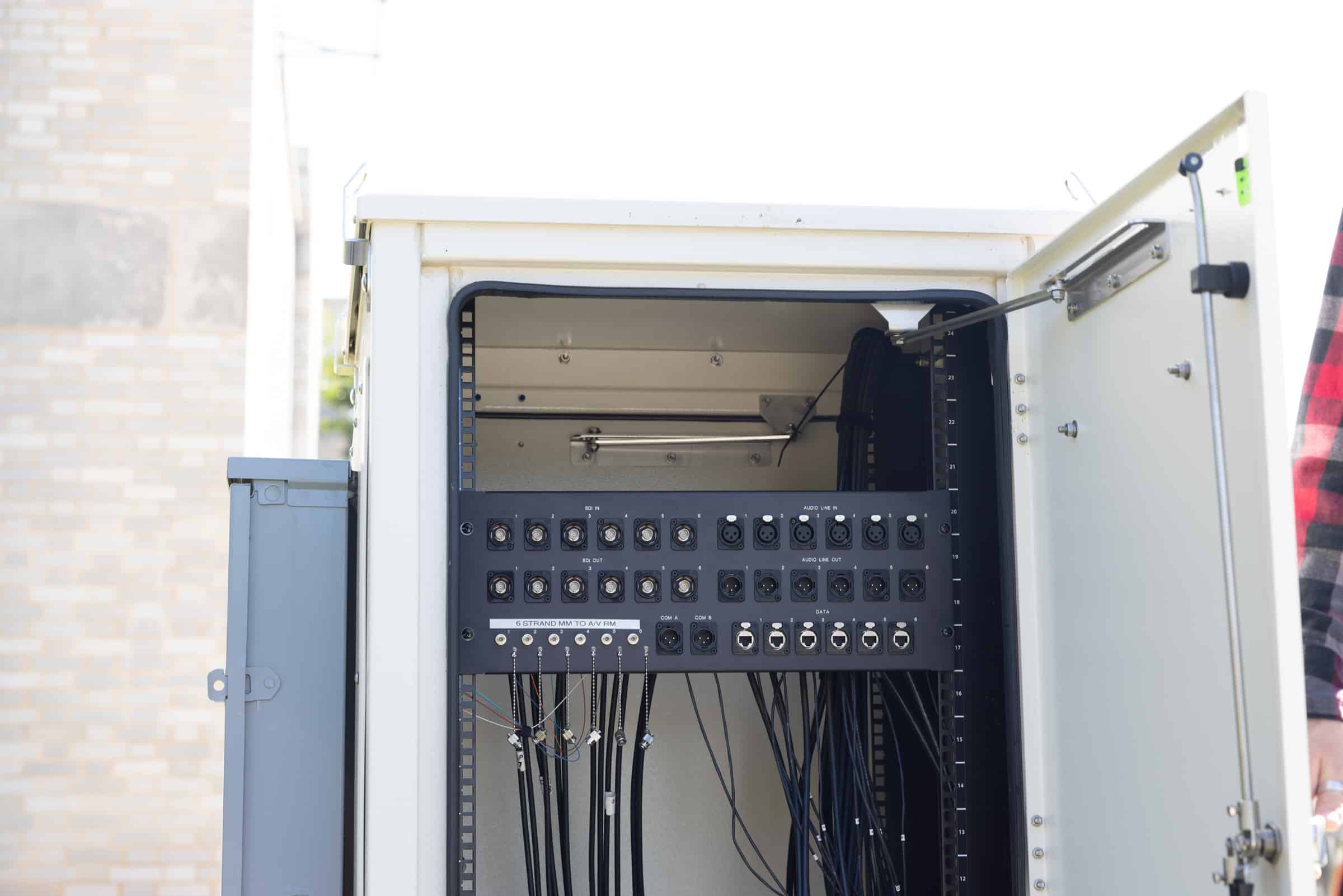 Open electrical cabinet with cables connected to ports, set outdoors by a brick wall, door revealing wiring inside.