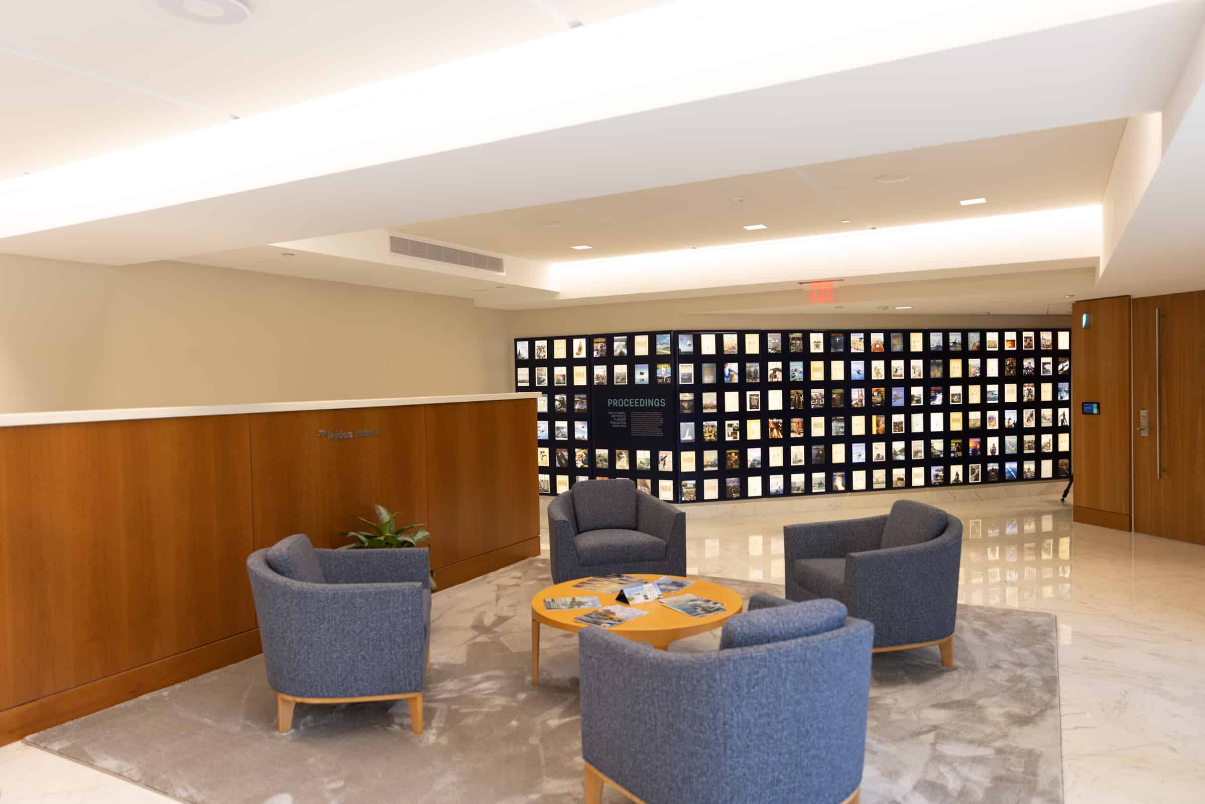 A modern lobby with marble floor, gray armchairs around a wooden table; magazine wall display and soft, welcoming lighting.