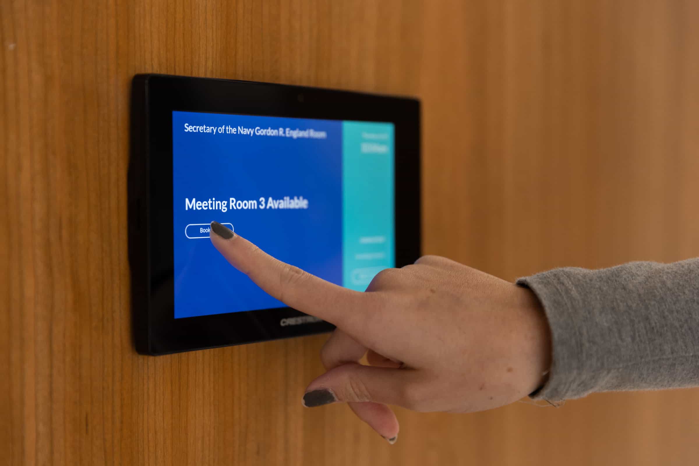 A hand with dark nail polish taps book on a touchscreen displaying Meeting Room 3 Available on a blue background.