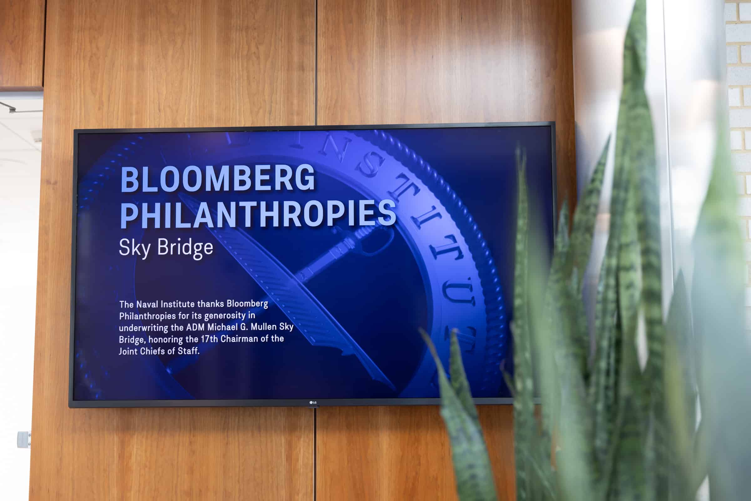 Digital screen thanks Bloomberg Philanthropies for underwriting Admiral Mullen Sky Bridge; plant visible in foreground.