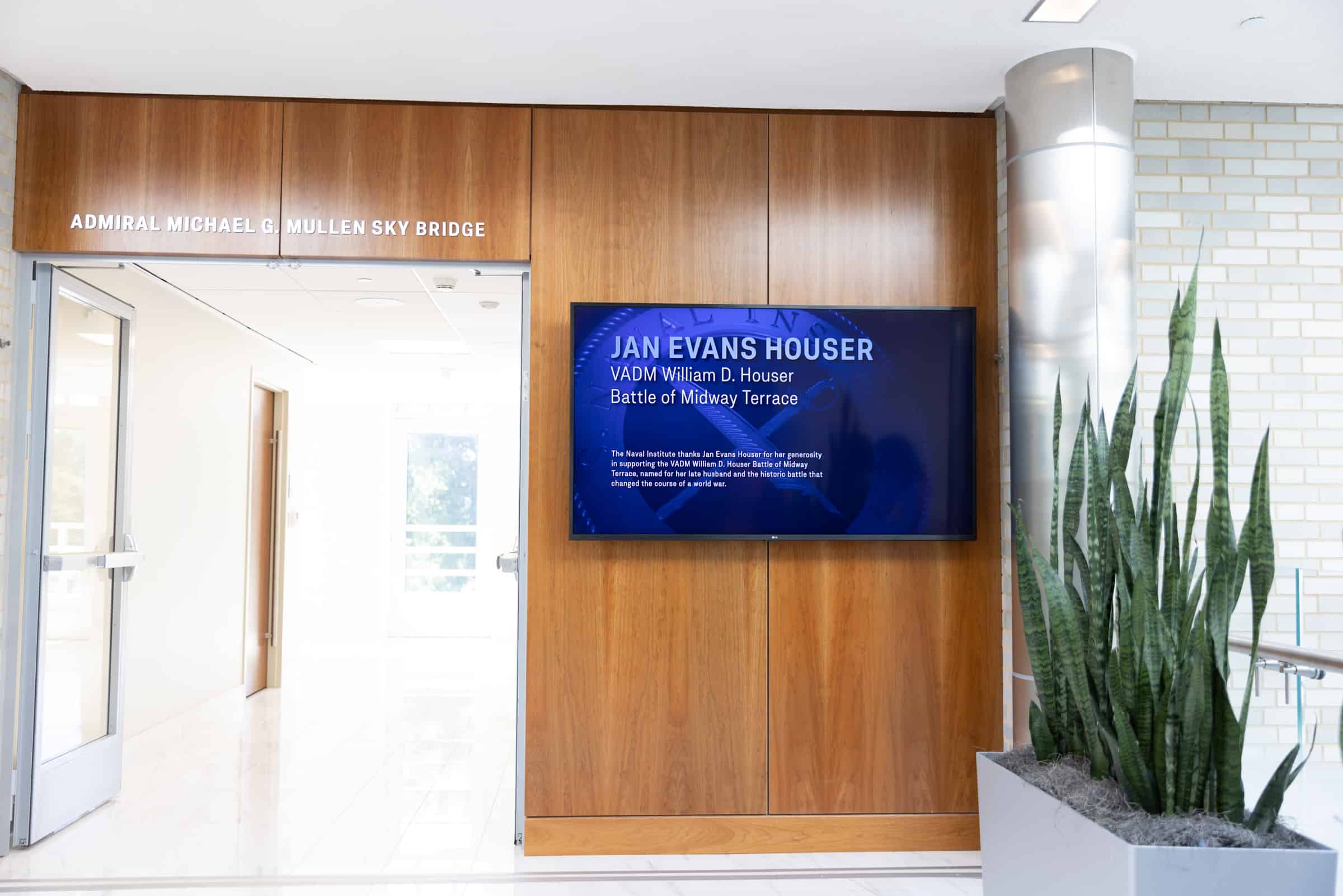 Hallway with wooden ADMIRAL MICHAEL G. MULLEN SKY BRIDGE sign, digital info screen, and a tall plant on the right.