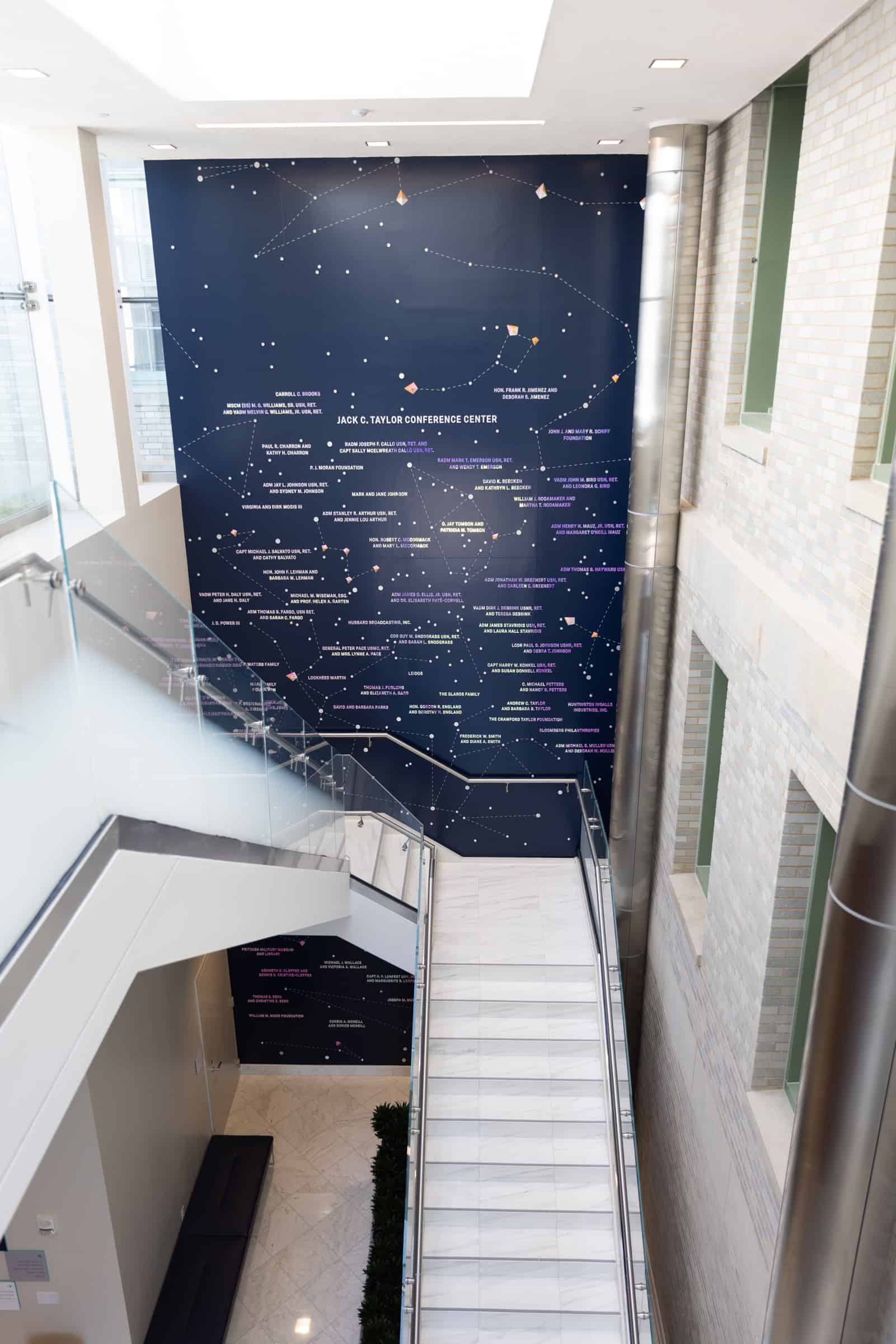 A modern conference center with a white staircase, featuring a dark blue constellation map wall under natural skylight illumination.